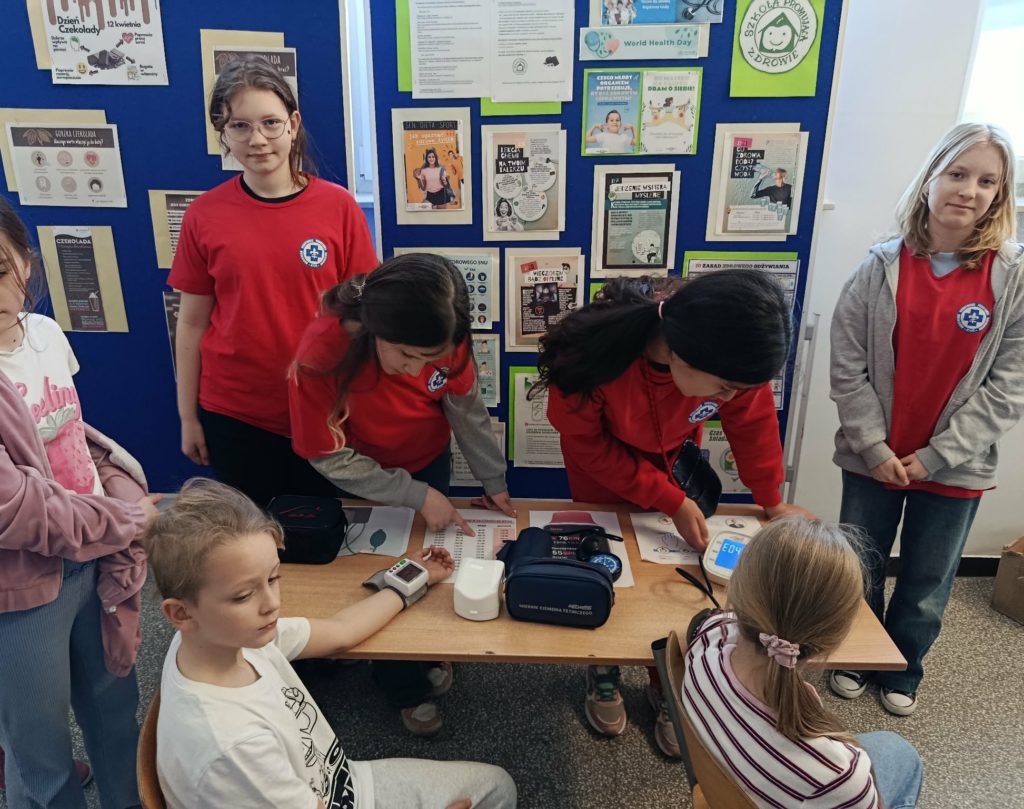 Przy stoliku na tle plakatów promujących zdrowy tryb życia i gorzką czekoladę stoją 4 uczennice Koła HOPR. Przed stolikiem trzech innych uczniów w czasie pomiaru ciśnienia tętniczego.