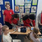 Przy stoliku na tle plakatów promujących zdrowy tryb życia i gorzką czekoladę stoją 4 uczennice Koła HOPR. Przed stolikiem trzech innych uczniów w czasie pomiaru ciśnienia tętniczego.