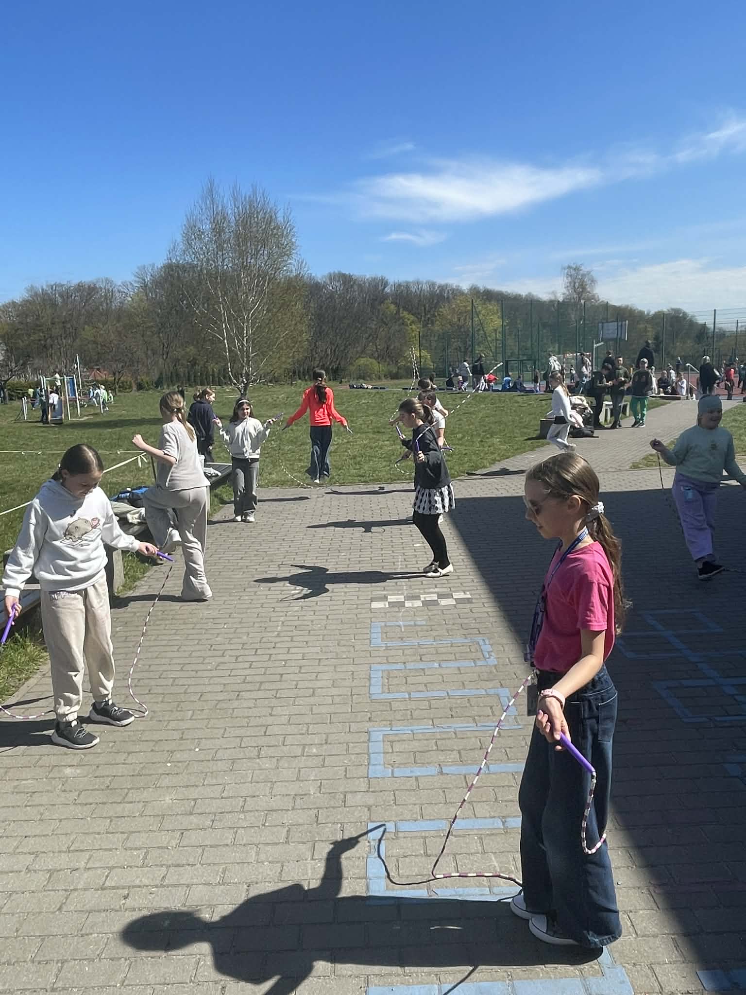 Grupa dzieci skacze na skakankach na brukowanej ścieżce na szkolnym podwórku w słoneczny dzień. Na pierwszym planie dziewczynka w różowej koszulce trzyma rączki fioletowej skakanki. W tle widać boisko i drzewa.