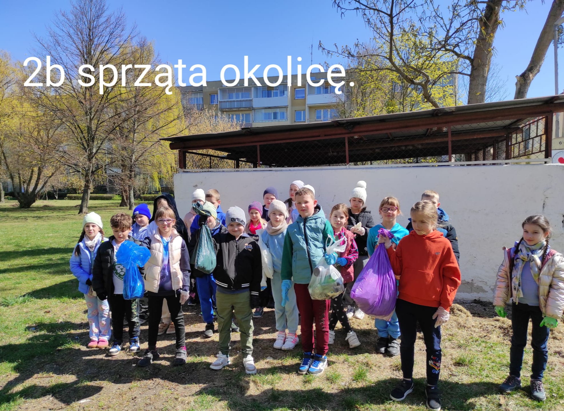 Uczniowie z klasy 2b stoją na placu przed szkołą.  Część z nich trzyma w dłoniach wypełnione worki na śmieci. U góry napis " 2B sprząta okolicę".