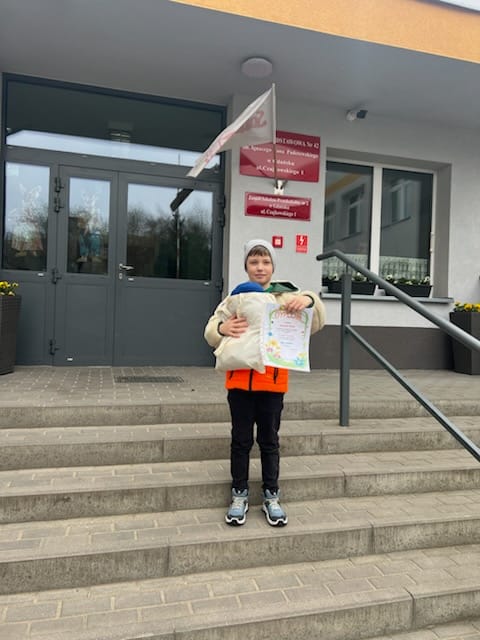 Uczeń stoi przed budynkiem szkoły na schodach, trzyma dyplom i nagrodę za konkurs.