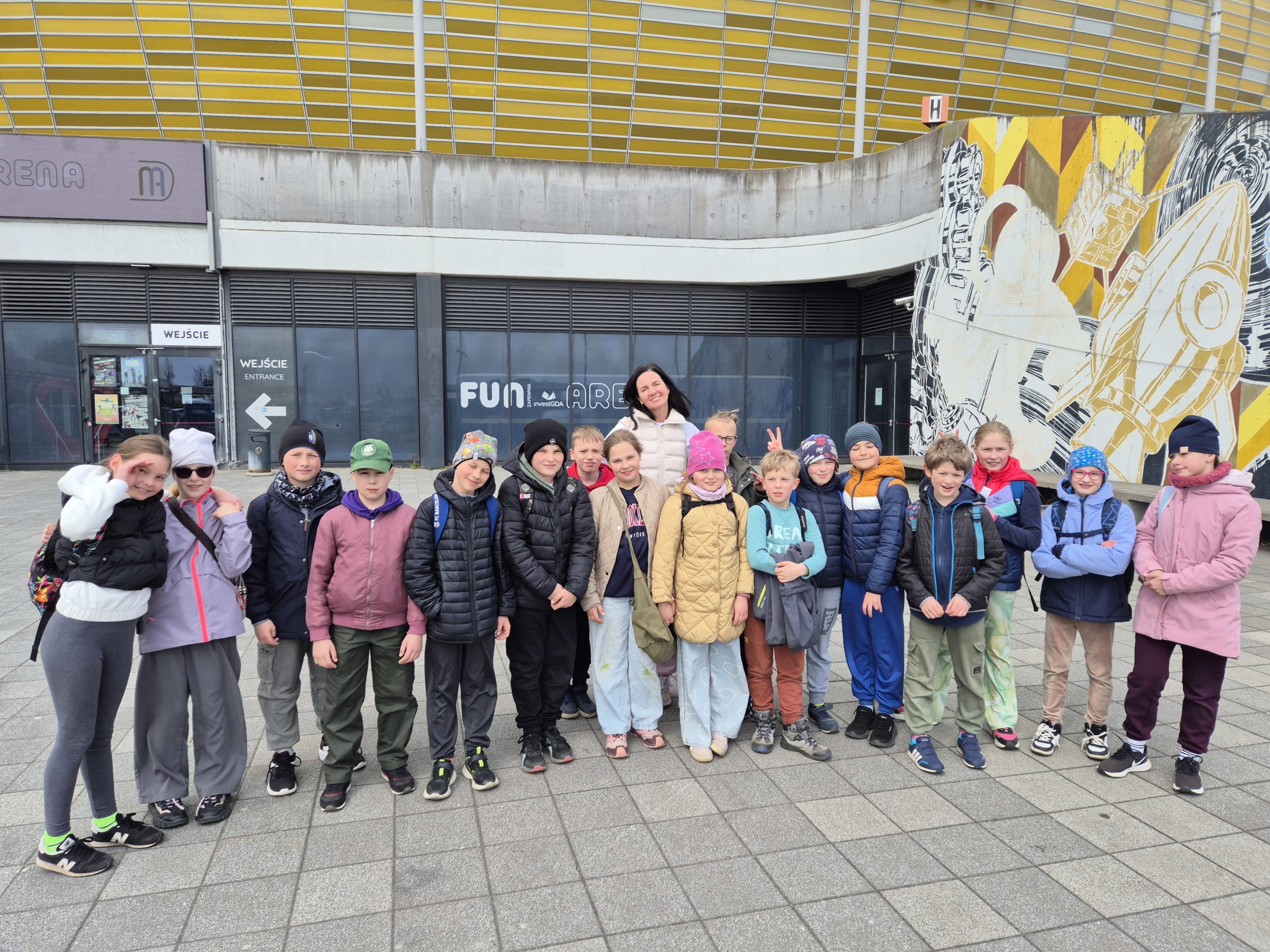 Widać grupę dzieci. Dzieci jest 17. Na środku jest z nimi Pani. Za dziećmi i panią widać napis Fun area. Po prawej stronie widać żółto białą ścianę.