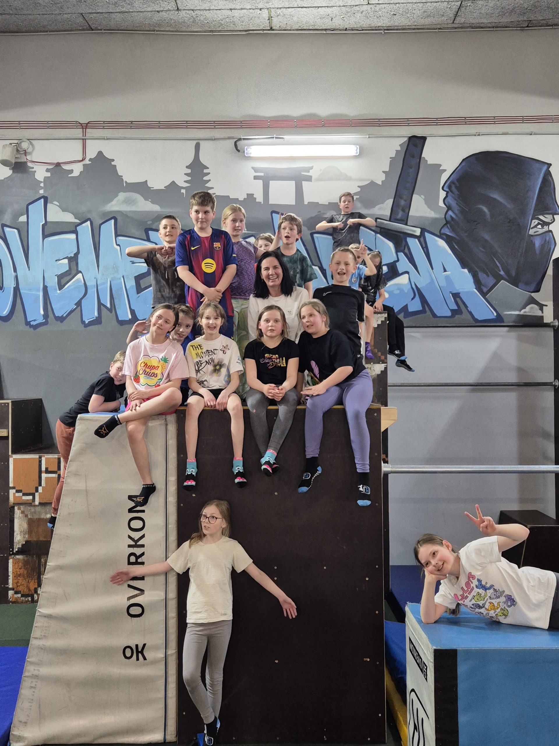 Na zdjęciu jest 17 dzieci. Na wysokim klocku znajdują się dzieci wraz z Panią. Część dzieci siedzi a część stoi. Na dole wysokiego klocka stoi jedna dziewczynka w okularach. Obok dużego klocka jest jeszcze kolejny klocek na nim jest jedna dziewczynka. Za dziećmi znajduje się napis MUVMENT ARENA.