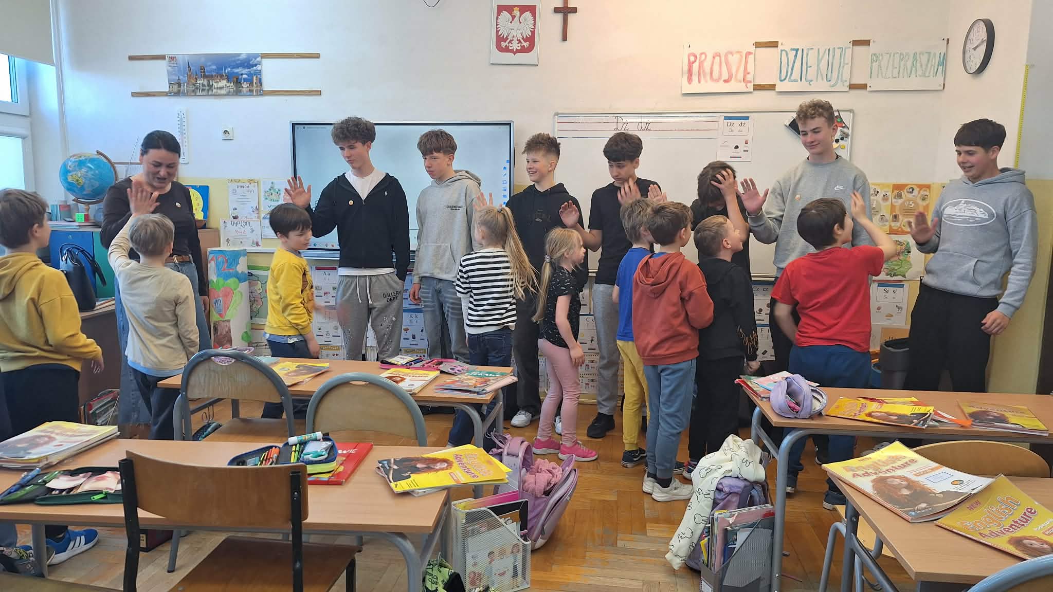 Grupa starszych i młodszych uczniów stoi w rzędach naprzeciw siebie w kolorowej sali lekcyjnej. Wyciągają do siebie dłonie w geście powitania lub zabawy („przybijanie piątki”). Na ścianie widoczne jest godło Polski oraz napisy „Proszę”, „Dziękuję”, „Przepraszam”.