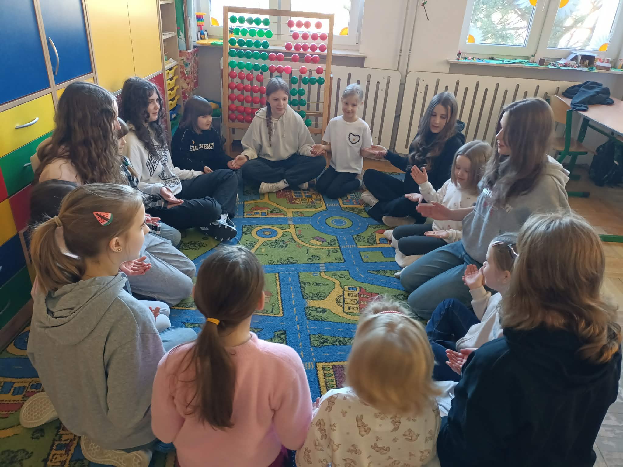 Grupa dzieci i młodzieży siedzi w kręgu na kolorowym dywanie z motywem ulic. Uczestnicy trzymają się za ręce lub wykonują wspólne gesty.