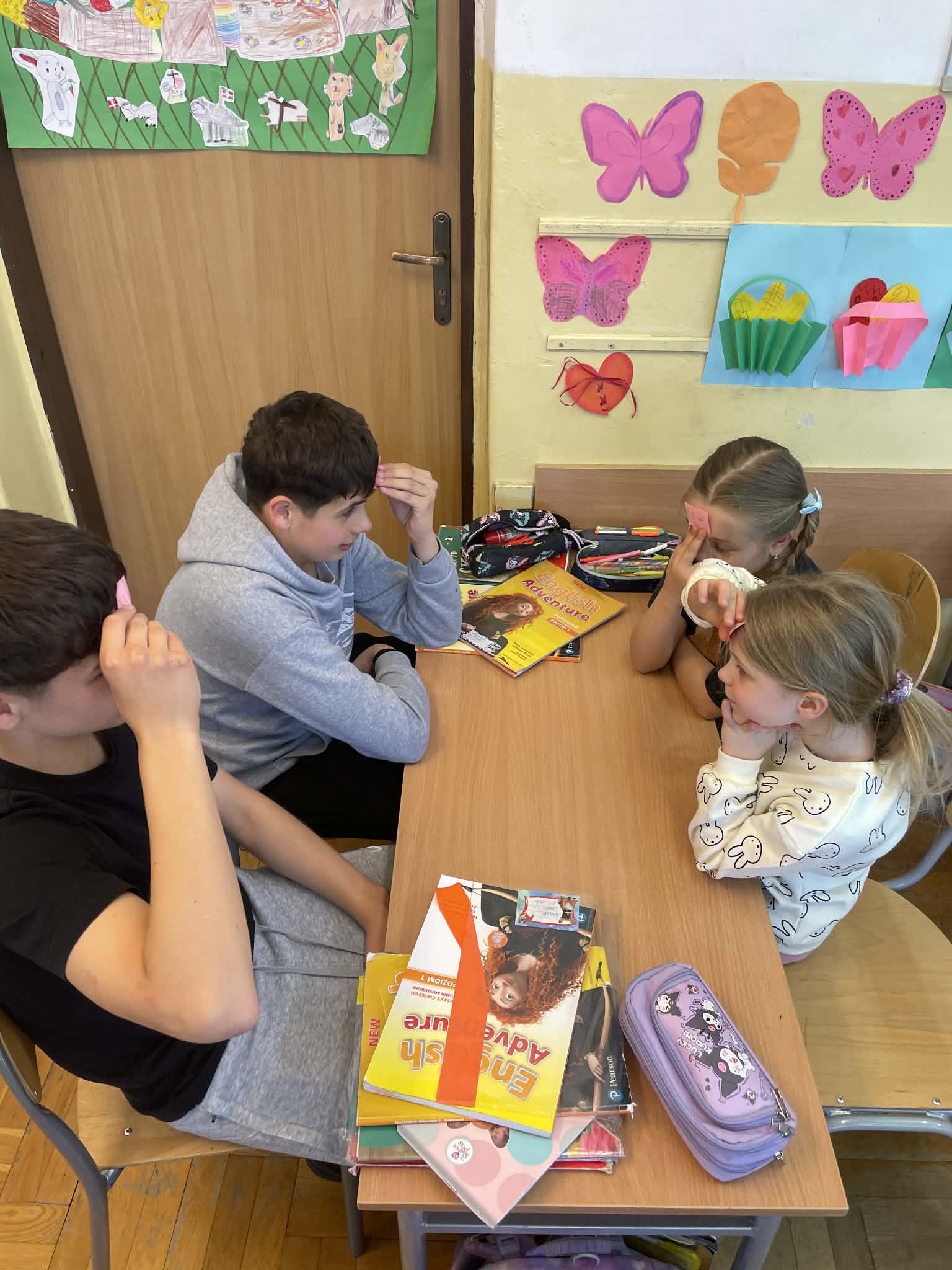 Czworo uczniów siedzi przy ławce, grając w grę polegającą na odgadywaniu haseł przyklejonych do czoła na kolorowych karteczkach. Na stole leżą podręczniki do języka angielskiego.