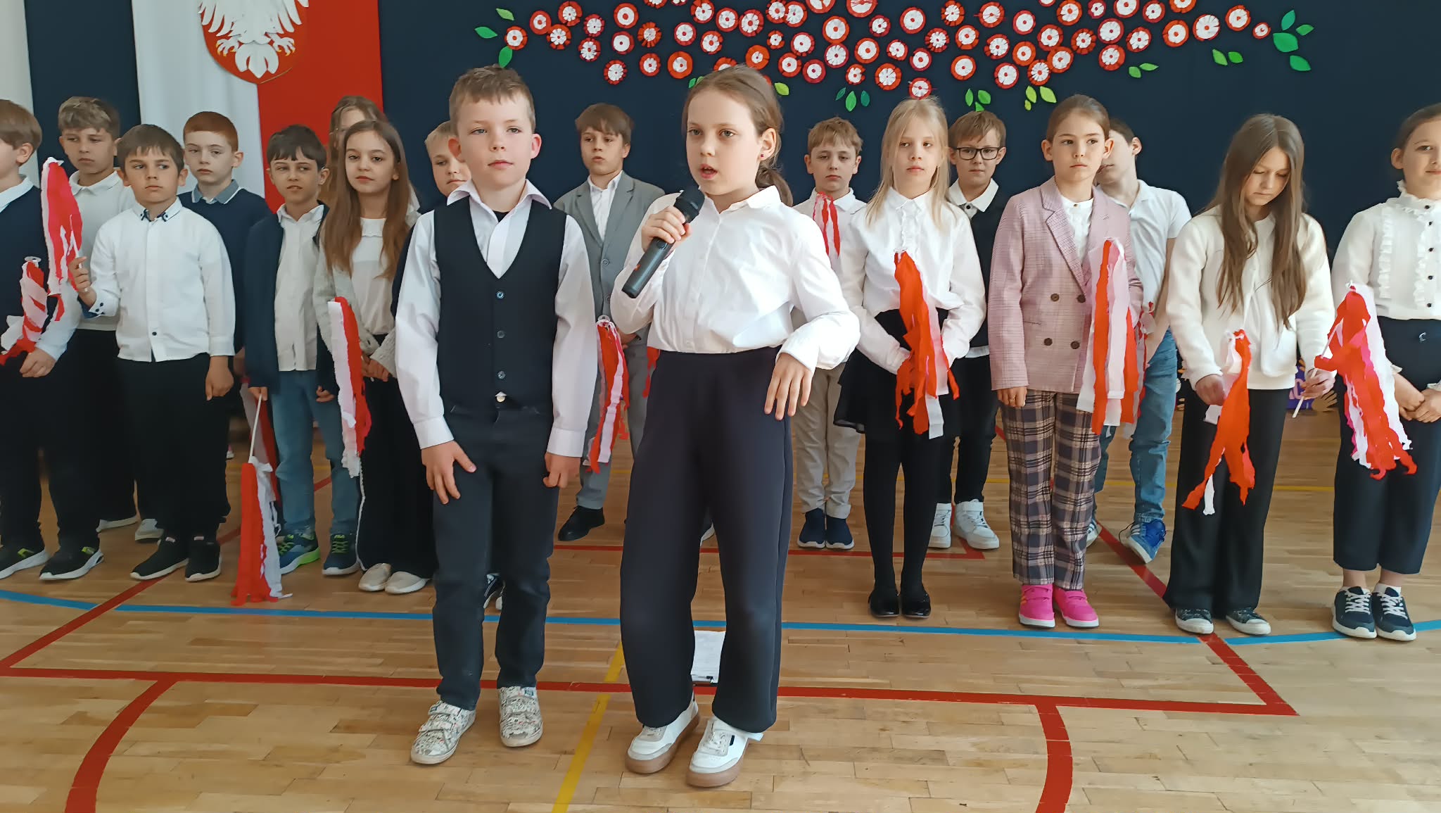 Dwoje uczniów stoi na środku i prezentuje część artystyczną z mikrofonem, za nimi grupa dzieci z rekwizytami w barwach narodowych.