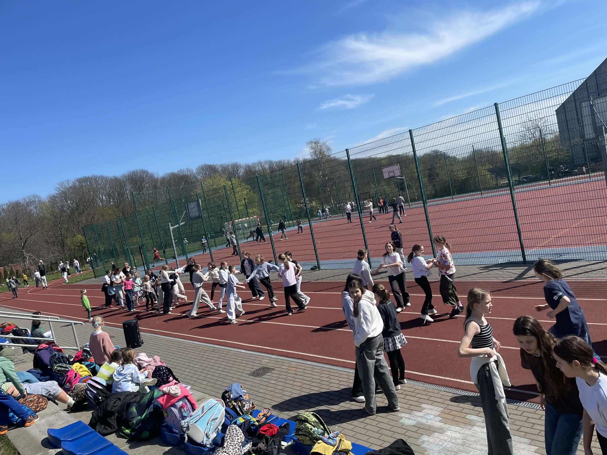Duża grupa uczniów tańczy belgijkę na czerwonej bieżni lekkoatletycznej na otwartym boisku. W tle widać zielone ogrodzenie, kosz do koszykówki i budynek szkoły. Pogoda jest bezchmurna.