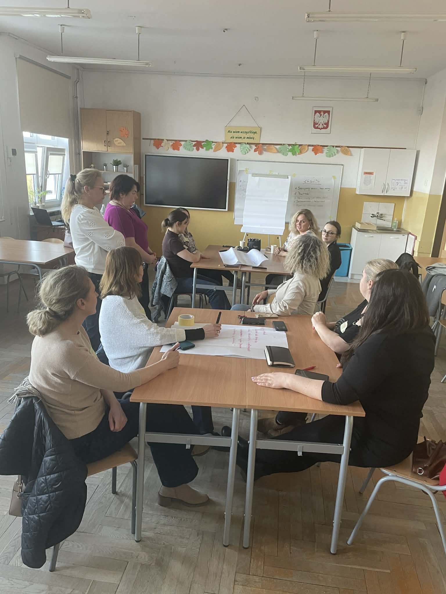 Grupa dorosłych osób pracuje w zespole przy szkolnej ławce. Część uczestników stoi, a część siedzi, omawiając zapiski na arkuszu papieru leżącym na stole. W tle widać tablicę z przypiętymi notatkami i ekran. 3