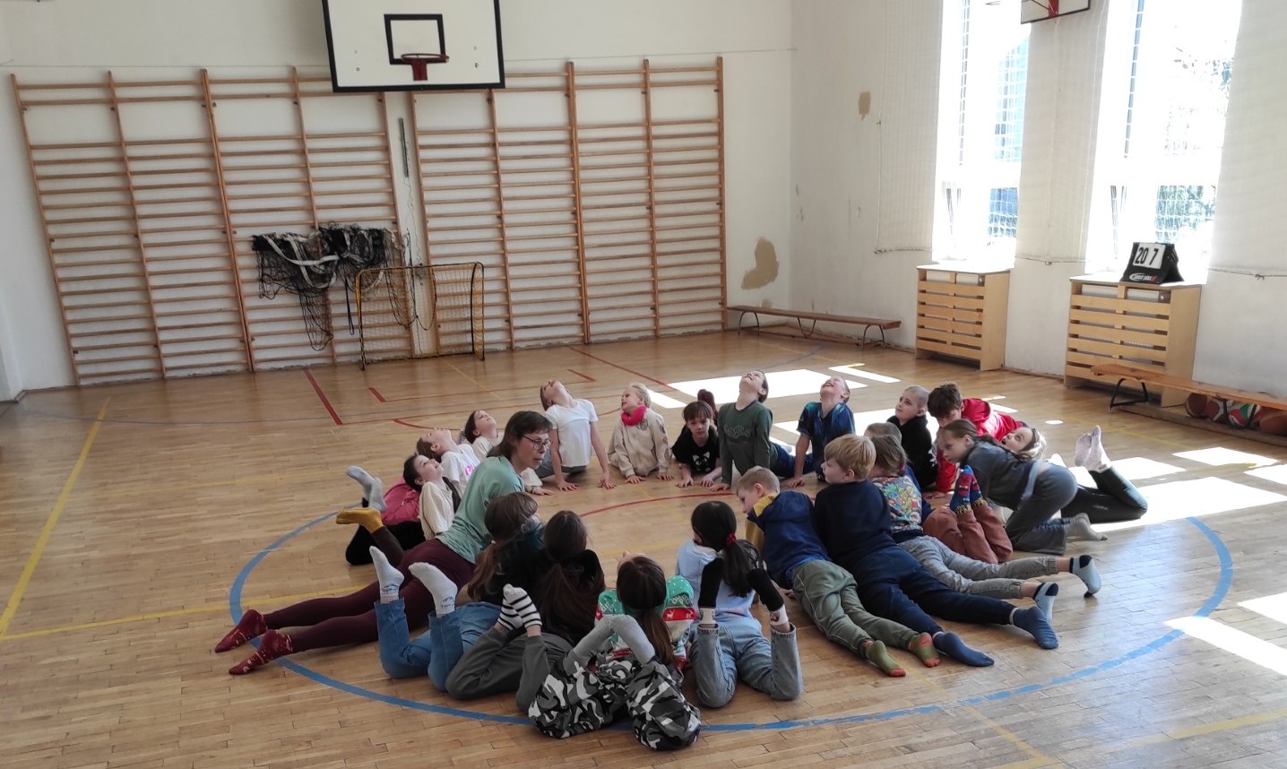 Grupa kilkunastu uczniów w różnobarwnych strojach sportowych leży na brzuchach w okręgu na drewnianym parkiecie sali gimnastycznej. Wszyscy mają uniesione głowy i klatki piersiowe, patrząc w górę i w bok, wykonując ćwiczenie rozciągające pod opieką instruktorki. W tle widać drabinki gimnastyczne i kosz do koszykówki.