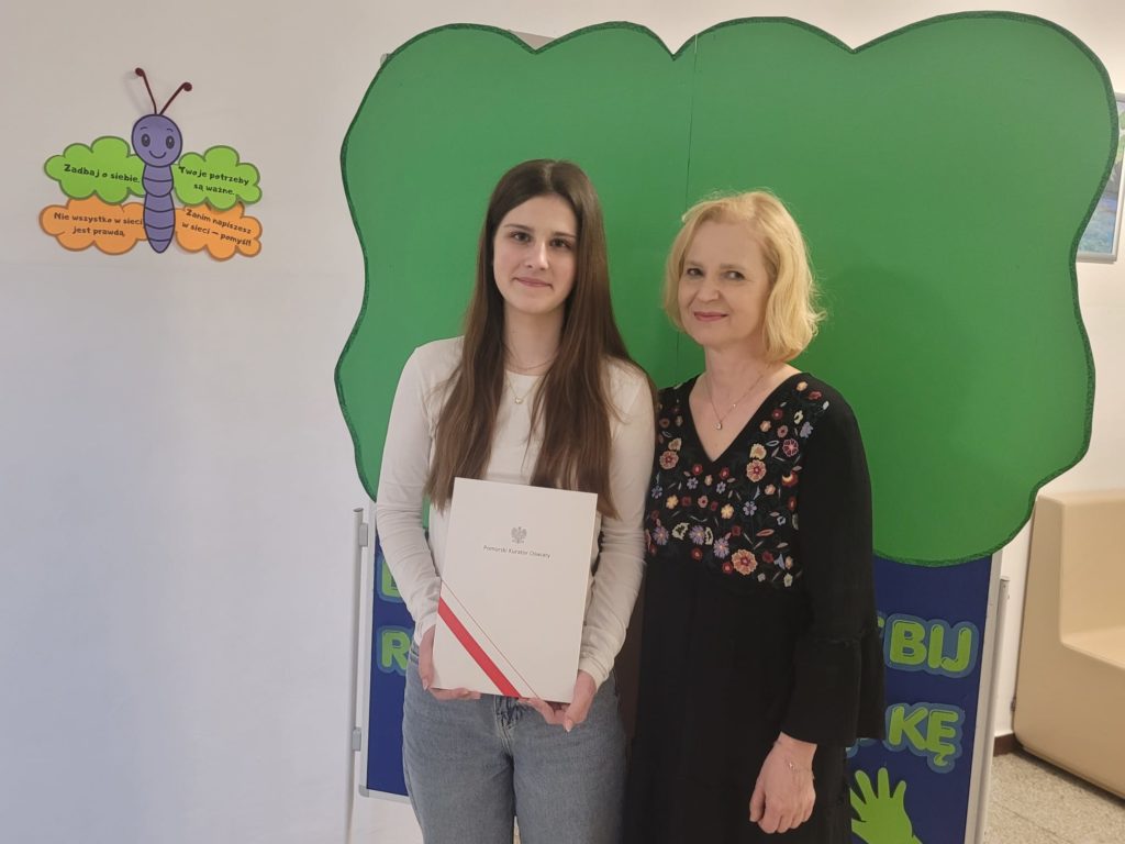 Małgosia stoi i trzyma w dłoniach dyplom. Obok stoi nauczycielka języka polskiego.