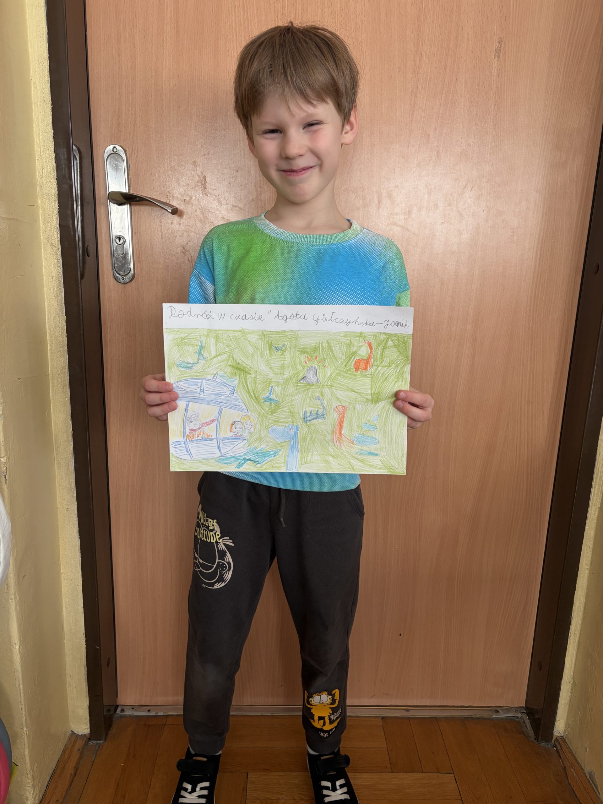 Chłopiec w niebieskiej bluzie sportowej stoi obok szkolnej tablicy, na której wisi duży błękitny plakat ze zdjęciami kapibar, a w ręku trzyma książkę zatytułowaną „Kapibara Barbara”.