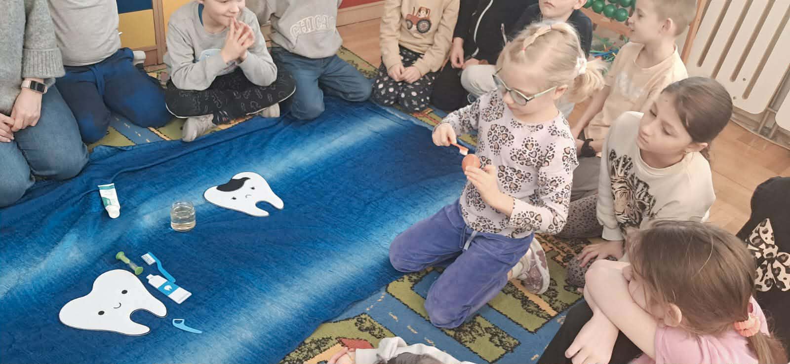 Grupa dzieci siedzi na dywanie w klasie, obserwując koleżankę, która demonstruje mycie zębów na modelu; wokół leżą szczoteczki, pasta i materiały edukacyjne przedstawiające zęby.