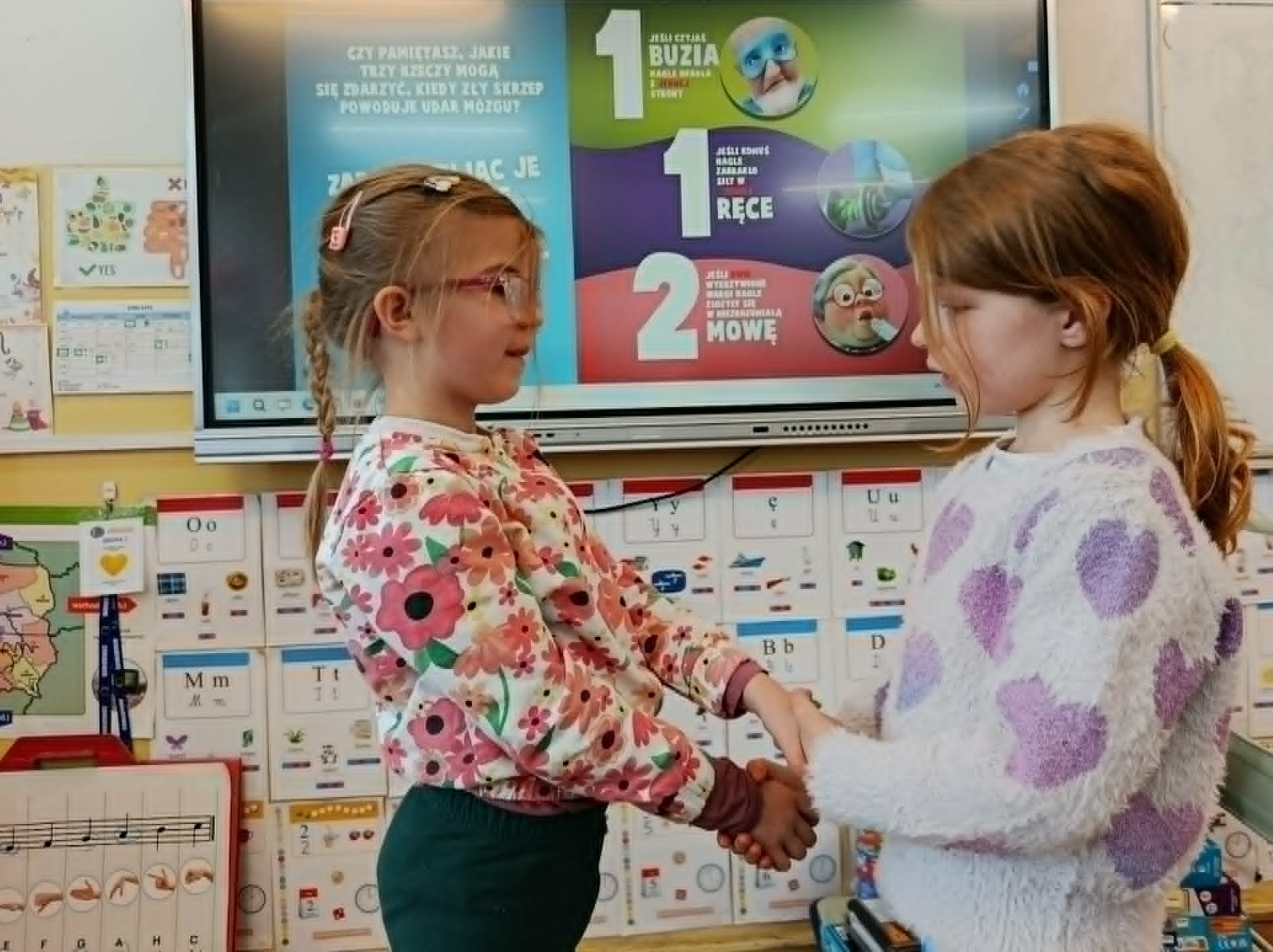 W sali lekcyjnej na tle tablicy multimedialnej z numerem 112 i objawami udaru mózgu stoją dwie dziewczynki trzymjace się za skrzyżowane ręce.