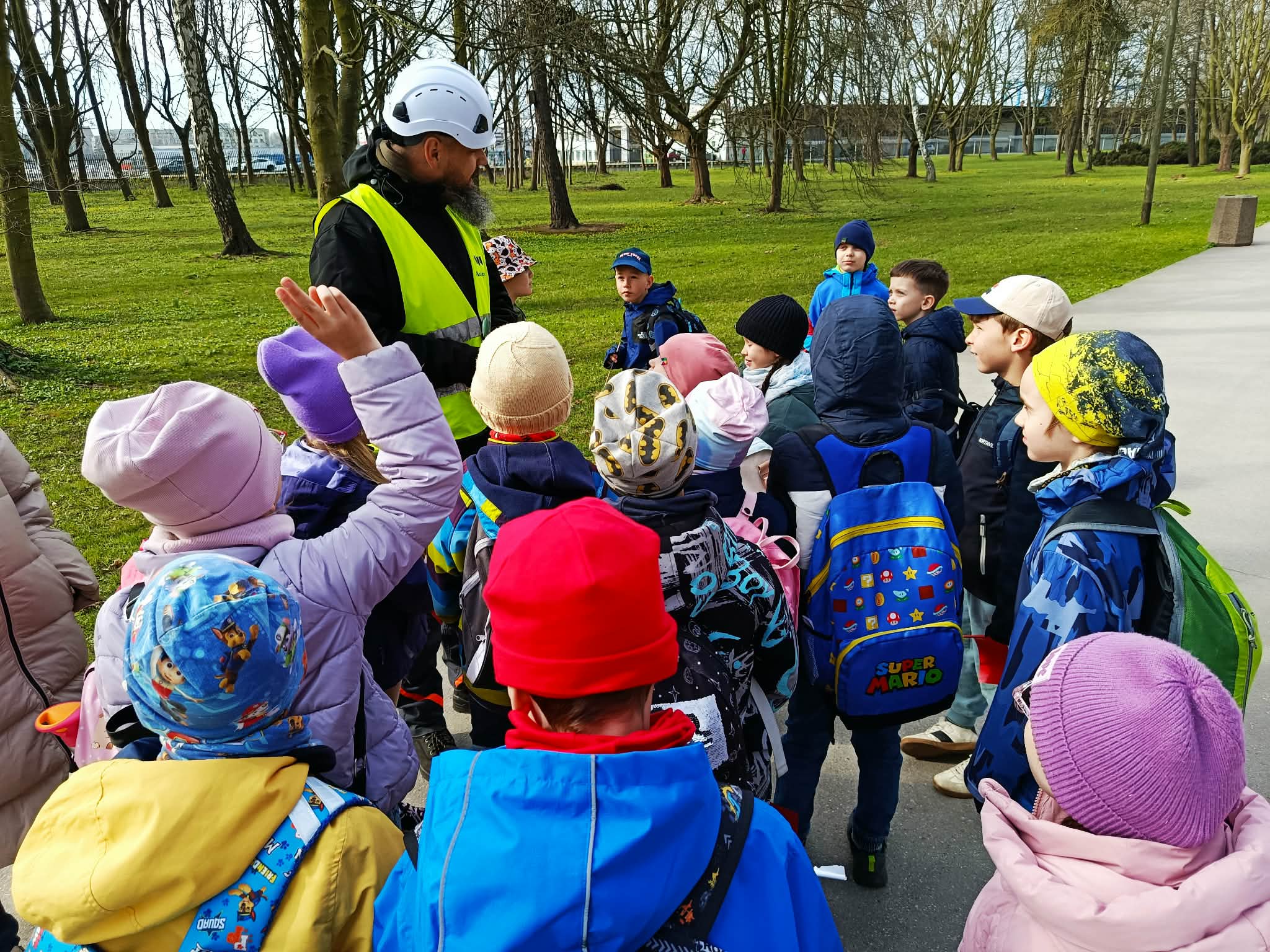 Grupa uczniów stojących w koło archeologa Muzeum Westerplatte. Jedna z dziewczynek podnosi rękę. W tle port.