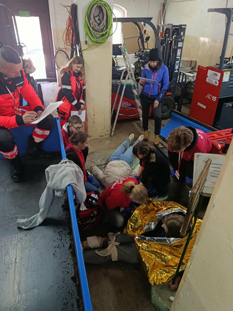 W garażu leży dwóch poszkodowanych pozorantów. W koło drużyna udzielająca pomocy i trzech ratowników oceniających ich działania.