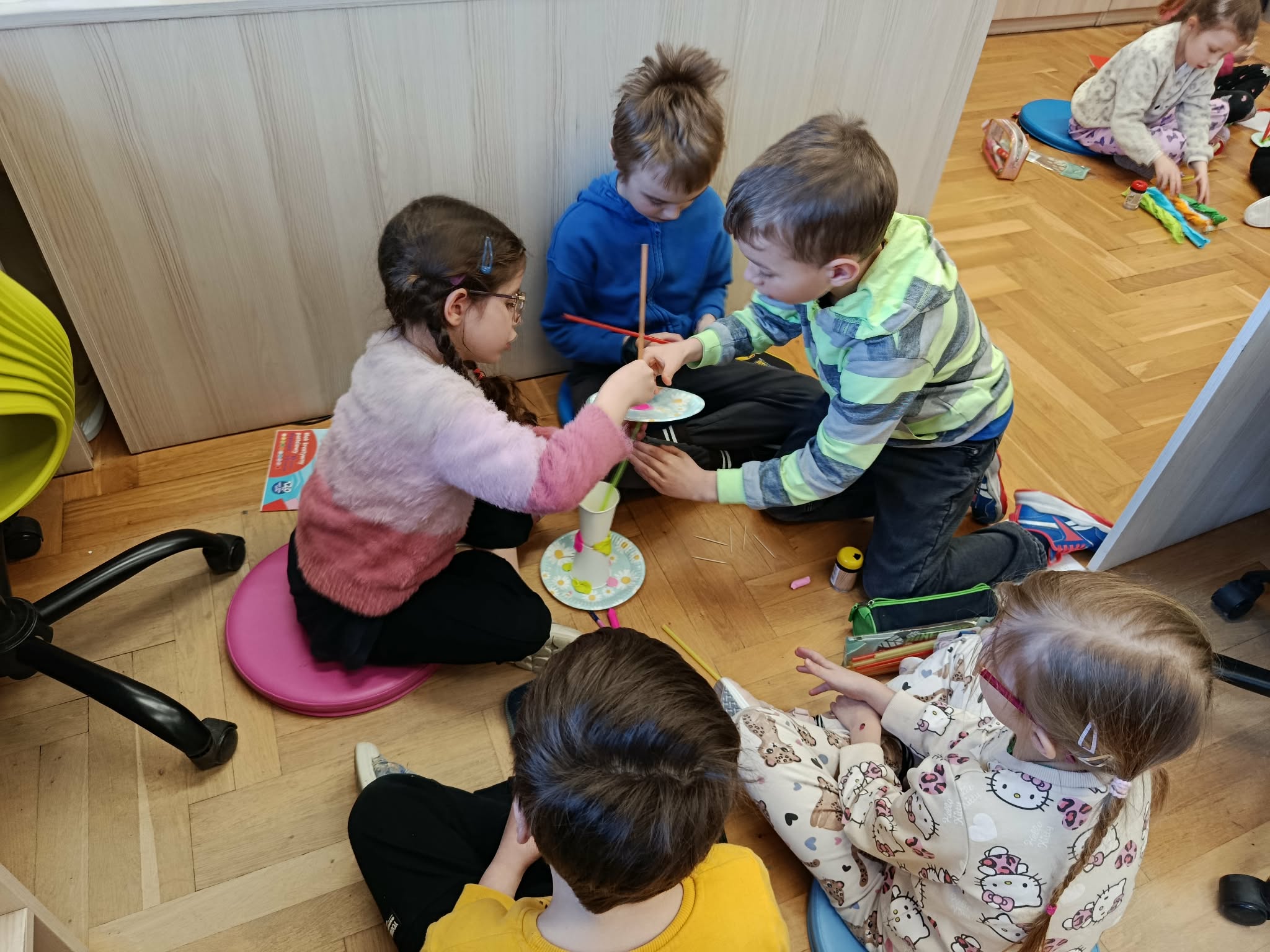 w kole na podłodze siedzi grupa dzieci i buduje konstrukcję ze słomek, talerzyków, ku-beczków, w koło leżą materiały.