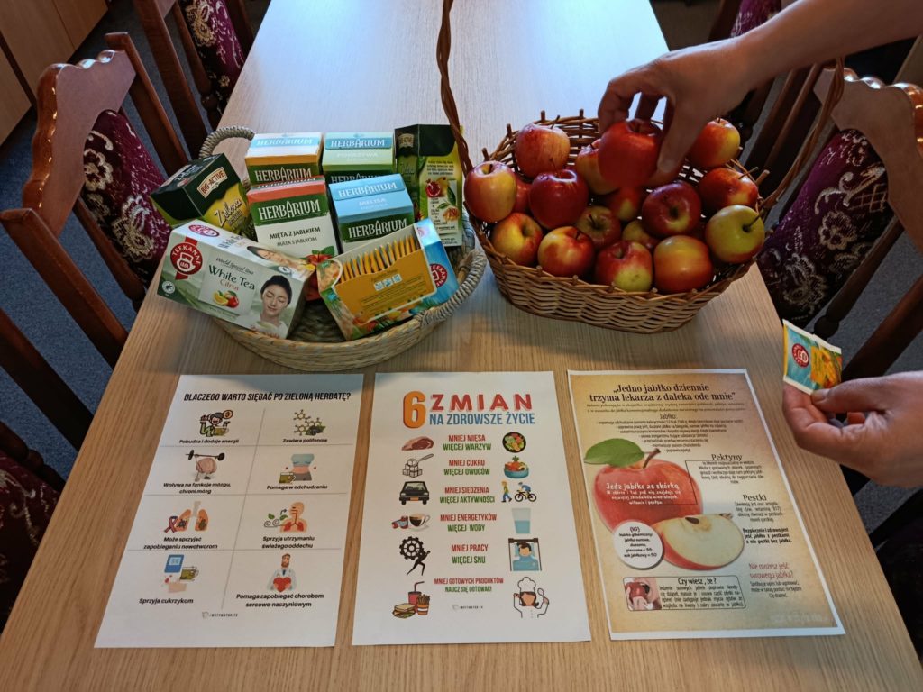 Na stole dwa wiklinowe kosze, w jednym z nich pudełka z ziołowymi herbatkami, w drugim jabłka i sięgająca po nie dłoń. Przed nimi plakaty na temat zdrowego trybu życia.