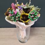 Ekologiczny bukiet wykonany z materiałów recyklingowych – kolorowe kwiaty z szyszek, papieru i innych elementów, ułożone w szklanym wazonie.