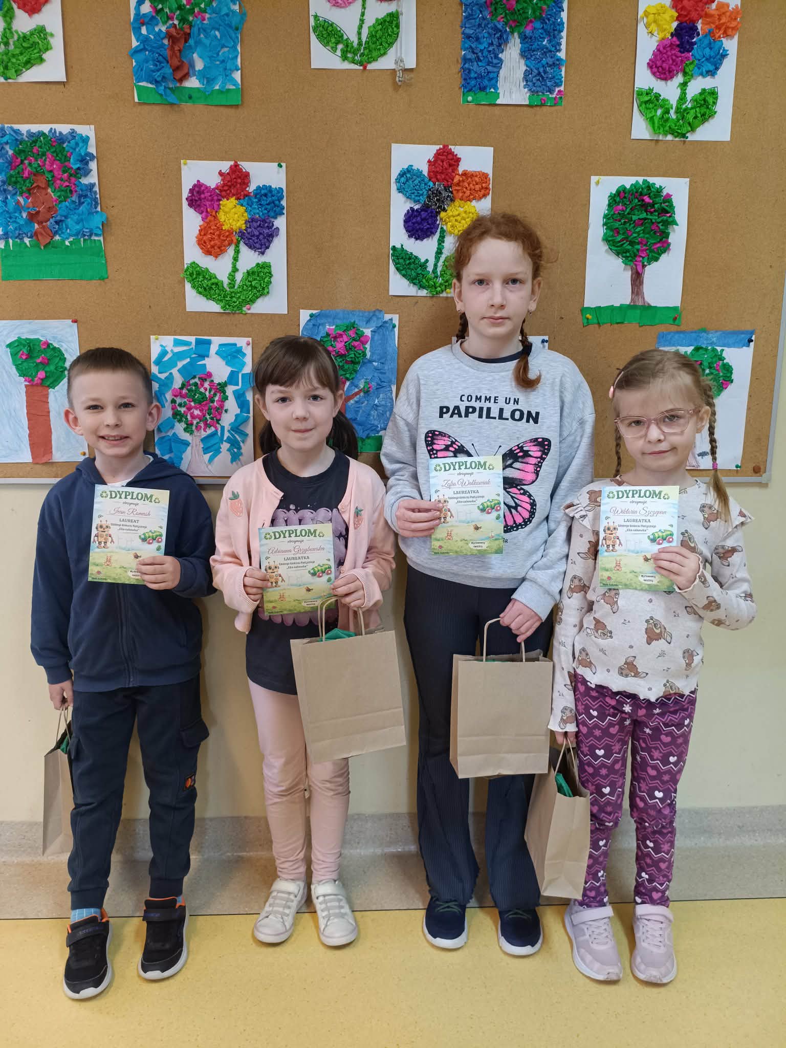 Czworo dzieci stoi w sali, trzymają dyplomy i papierowe torby, na tle tablicy z pracami plastycznymi przedstawiającymi kwiaty.