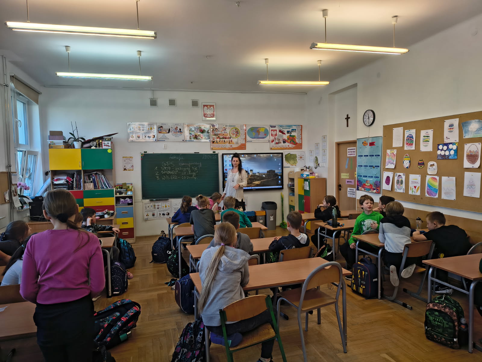 W sali lekcyjnej grupa uczniów szkoły podstawowej siedzi przy ławkach tyłem do obiektywu, słuchając nauczycielki stojącej przy tablicy. Na zielonej tablicy widać zapisane hasła i datę, obok znajduje się ekran multimedialny. Ściana jest ozdobiona pracami plastycznymi uczniów.