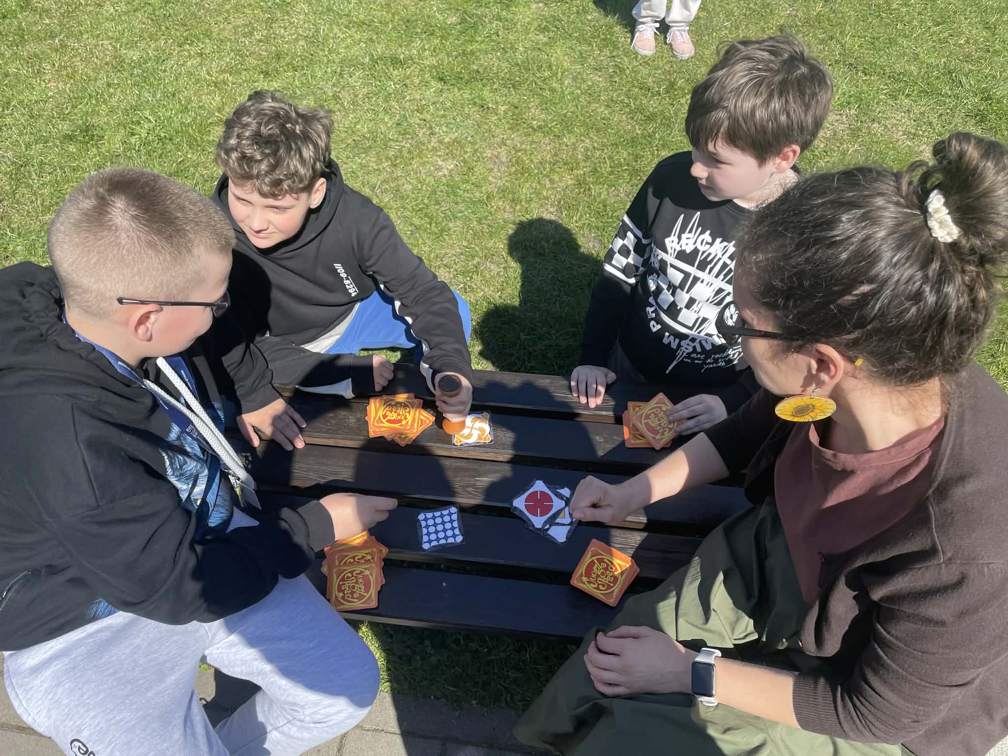 Troje chłopców i kobieta siedzą przy drewnianym stole piknikowym na trawie i grają w grę karcianą. Chłopiec w okularach i kobieta patrzą na karty rozłożone na stole.
