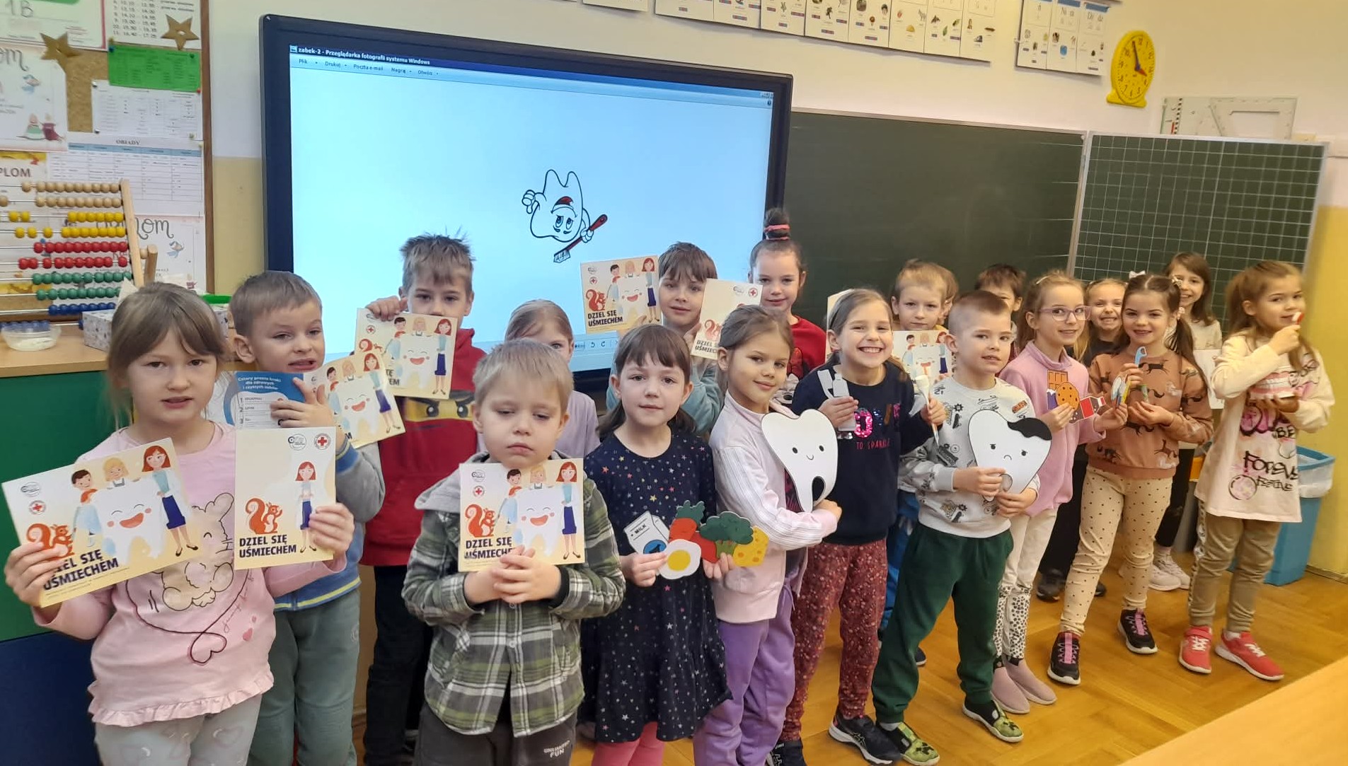 Trochę bardziej szczegółowy: Dzieci stoją w rzędzie w sali lekcyjnej, trzymając prace i pomoce dotyczące dbania o zęby; w tle tablica i ekran z ilustracją zęba.