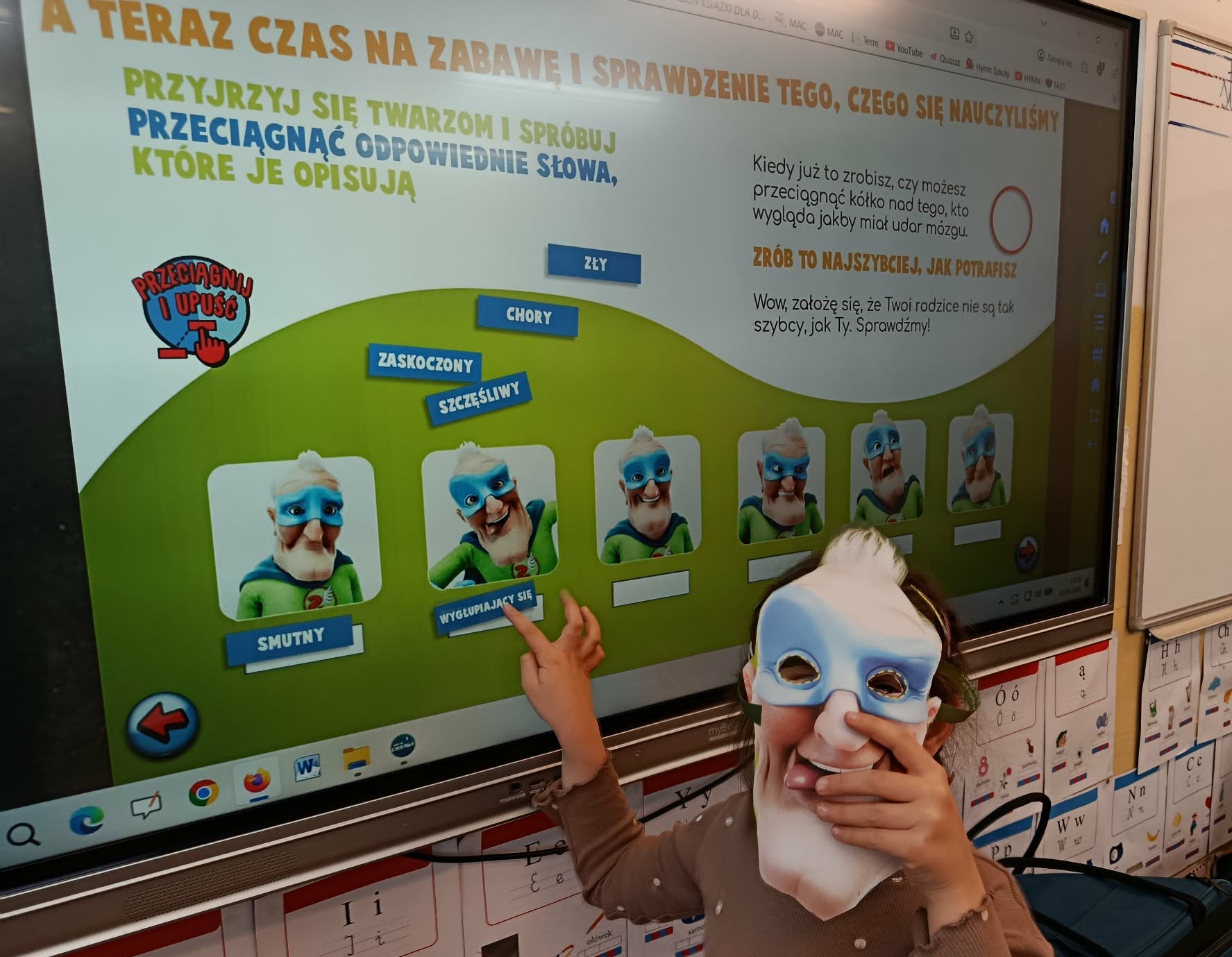 Na tle tablicy multimedialnej wyświetlającej różne miny stoi dziewczynka z maską dziadka na twarzy.