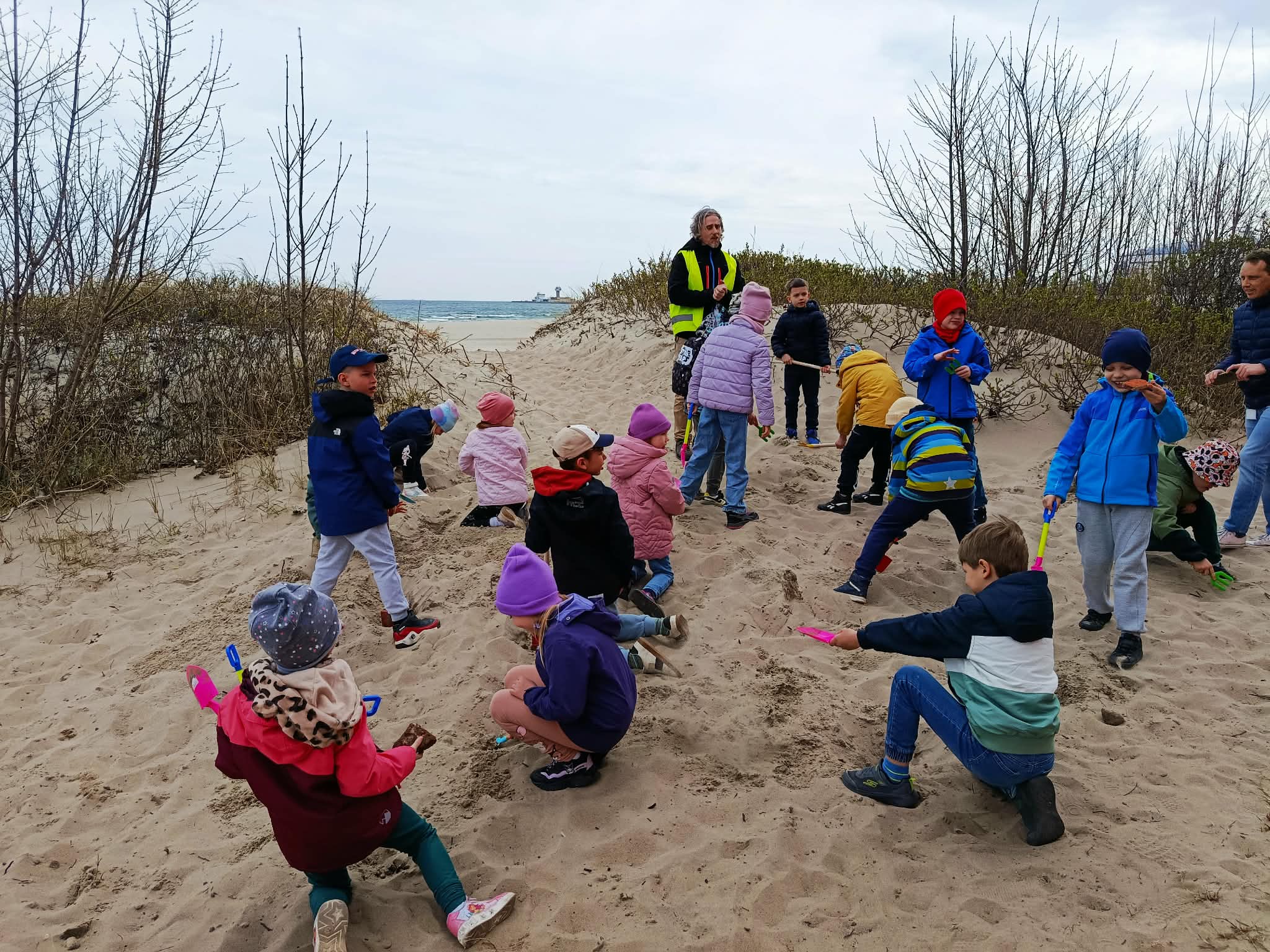 Grupa uczniów kopiących łopatkami w plażowym piasku na wydmie, przed nimi przewod-nik. W tle morze.