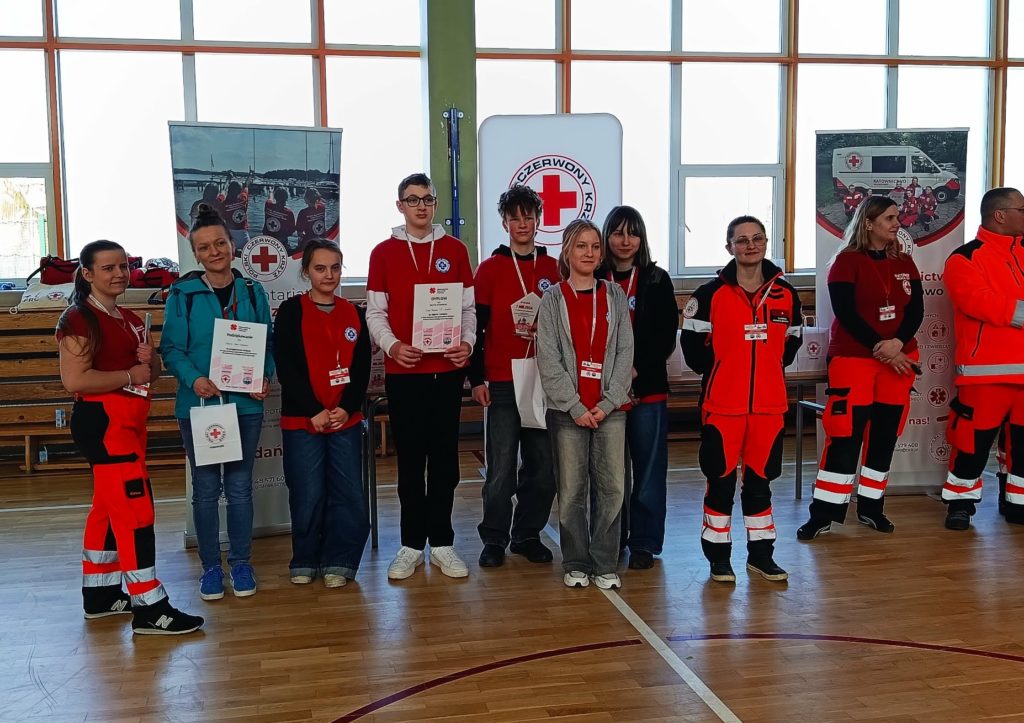 Na tle banerów PCK drużyna SP 47 w Gdańsku z opiekunem odbierający nagrodę za zajęcie I miejsca w etapie rejonowym XXXII Ogólnopolskich Mistrzostw Pierwszej Pomocy PCK. Obok organizatorzy i ratownicy.