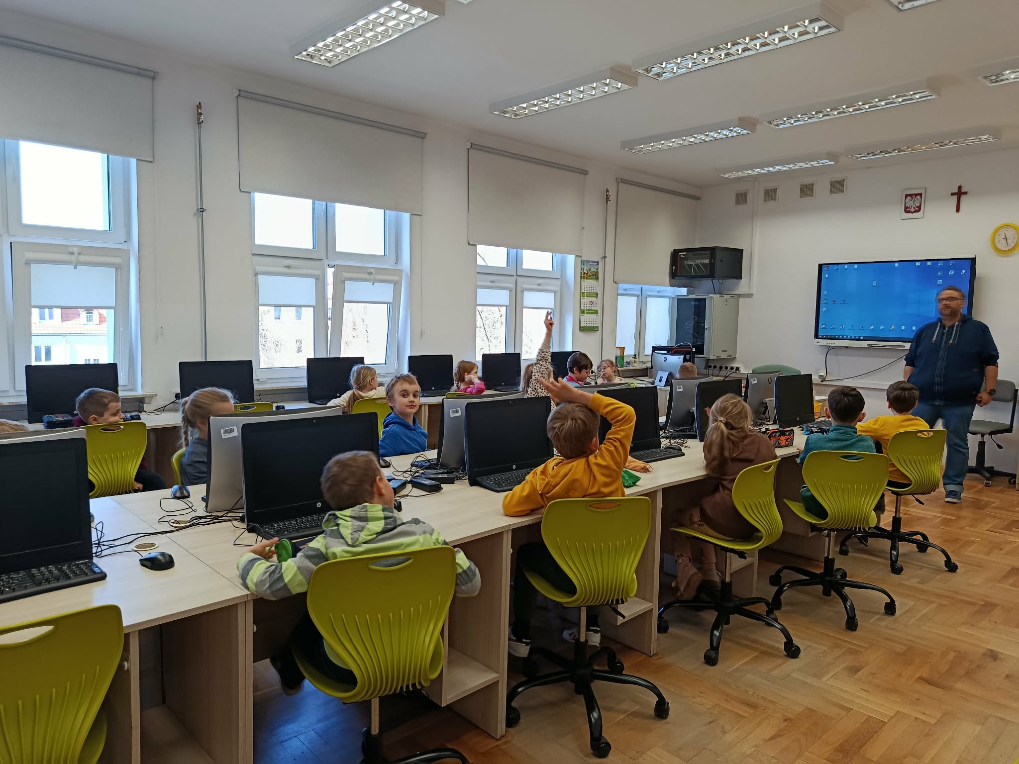 W sali komputerowej przed komputerami siedzą uczniowie klasy 1c. W tle przed tablicą stoi mężczyzna