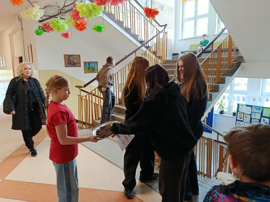 Na szkolnym korytarzu stoi uczennica Koła PP trzymająca tacę z gorzką czekoladą. Za nią nauczyciela, przed nią grupa uczennic częstujących się czekoladą.