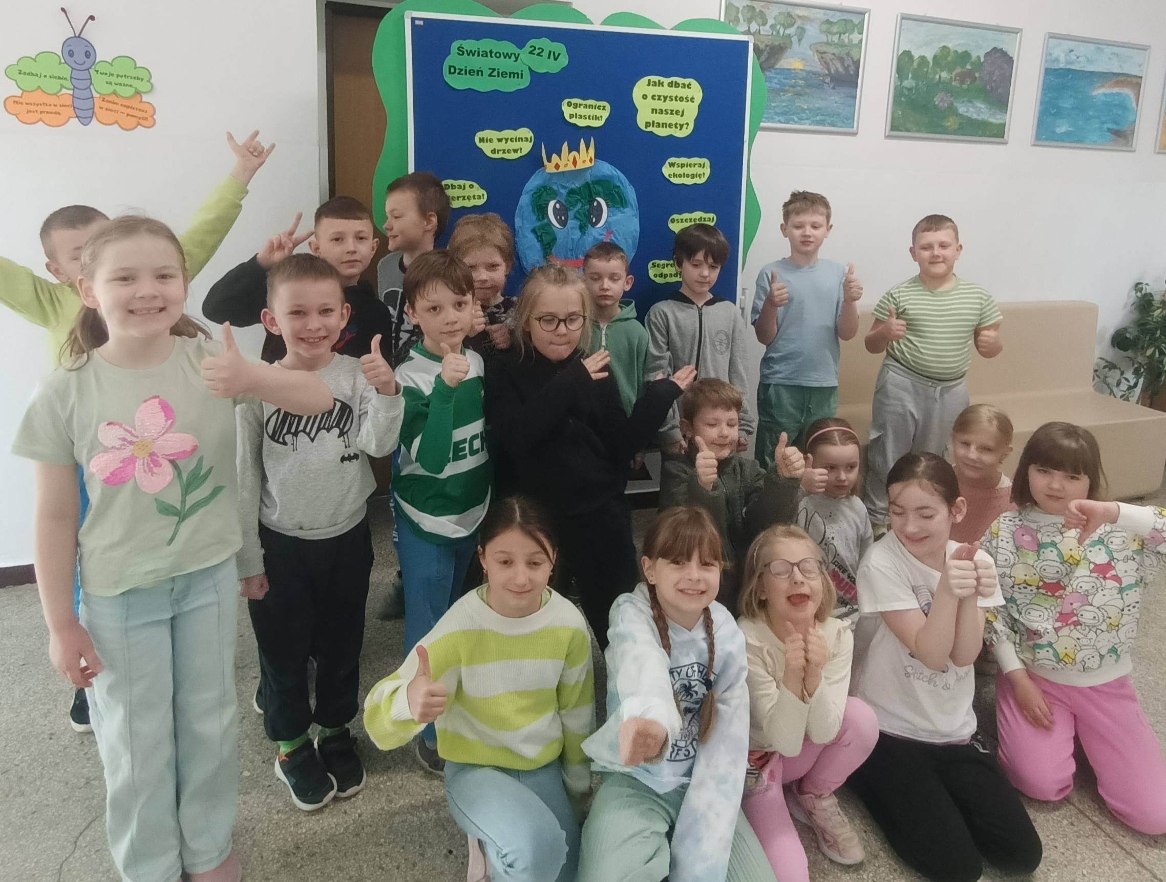 Grupa dzieci ustawiona w trzech rzędach pozuje na tle tablicy przedstawiającej kulę ziemską w koronie. Wokół kuli umieszczono hasła nawołujące do dbania o naszą planetę, np. " Segreguj odpady", "Oszczędzaj wodę" i "Dbaj o zwierzęta".