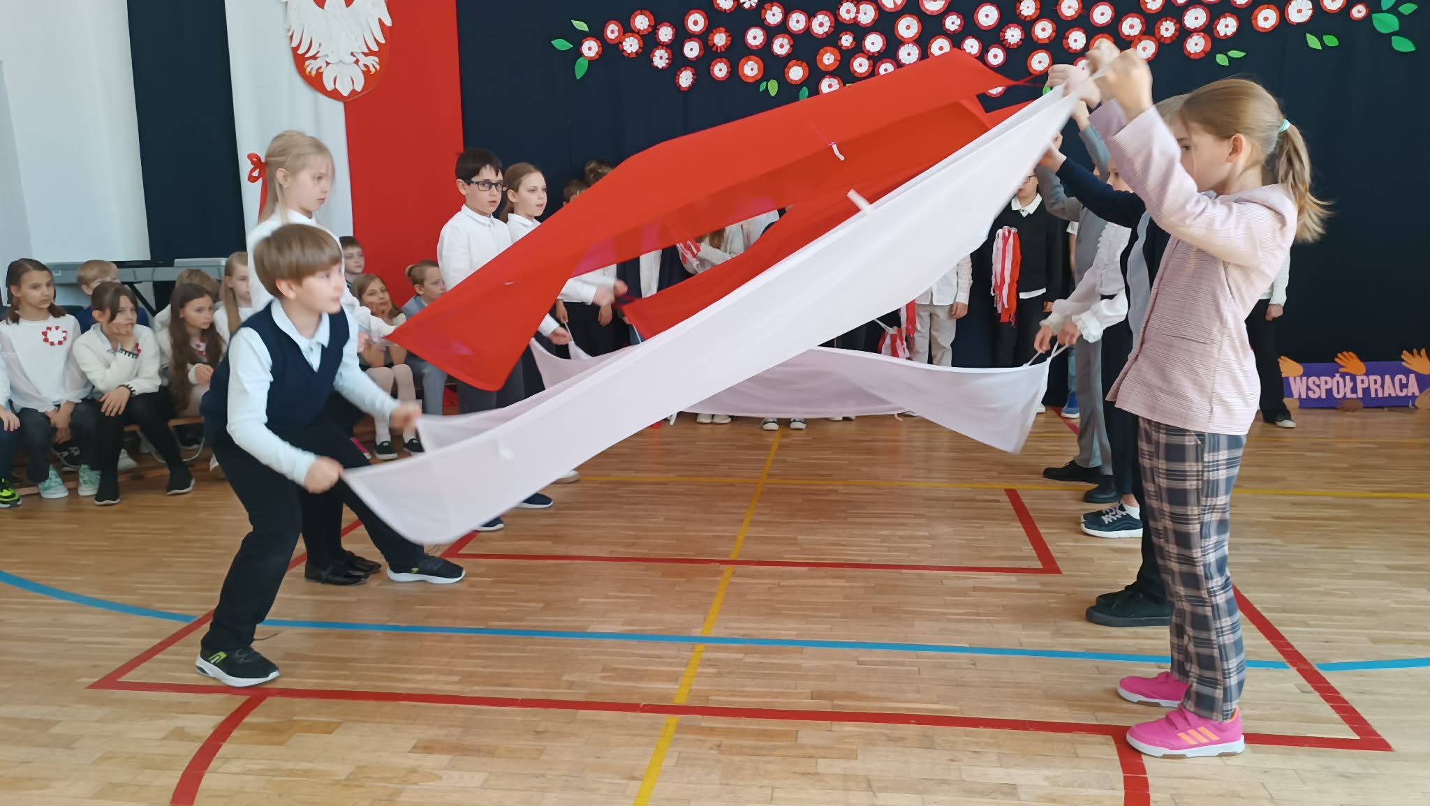 Dzieci w szkolnej sali gimnastycznej trzymają biało-czerwoną tkaninę, unosząc ją w powietrzu podczas występu. W tle siedzą inni uczniowie, a ściana udekorowana jest symbolami narodowymi.