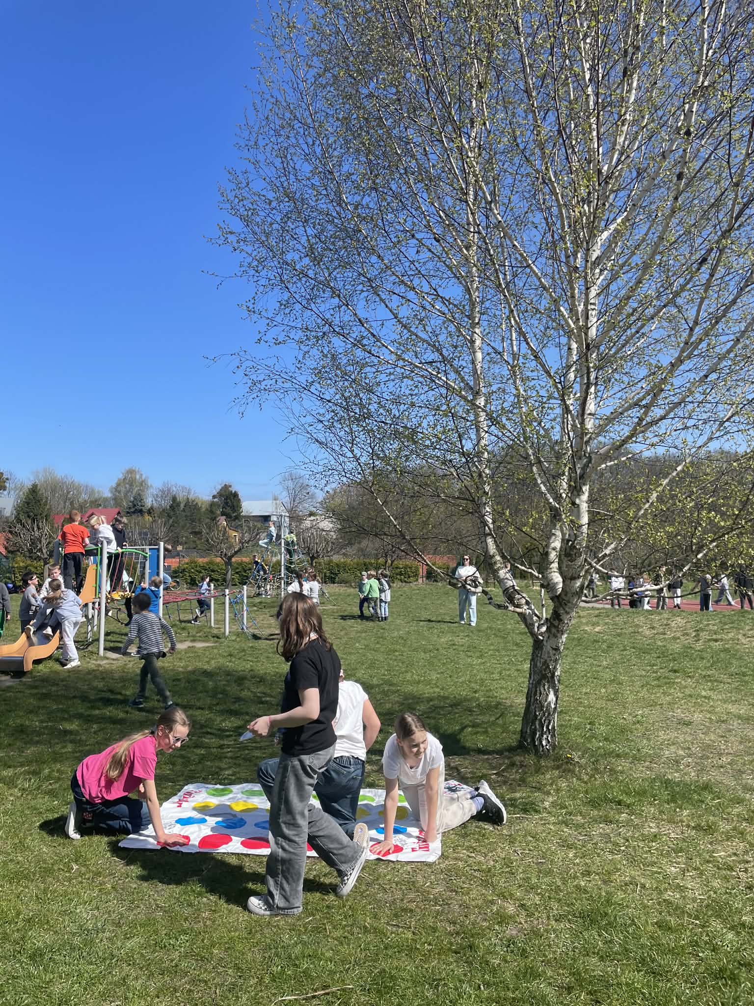 Trzy dziewczynki grają w Twistera na trawie, na białej macie z kolorowymi kołami. W tle widać inne dzieci bawiące się na placu zabaw z drabinkami i zjeżdżalnią.