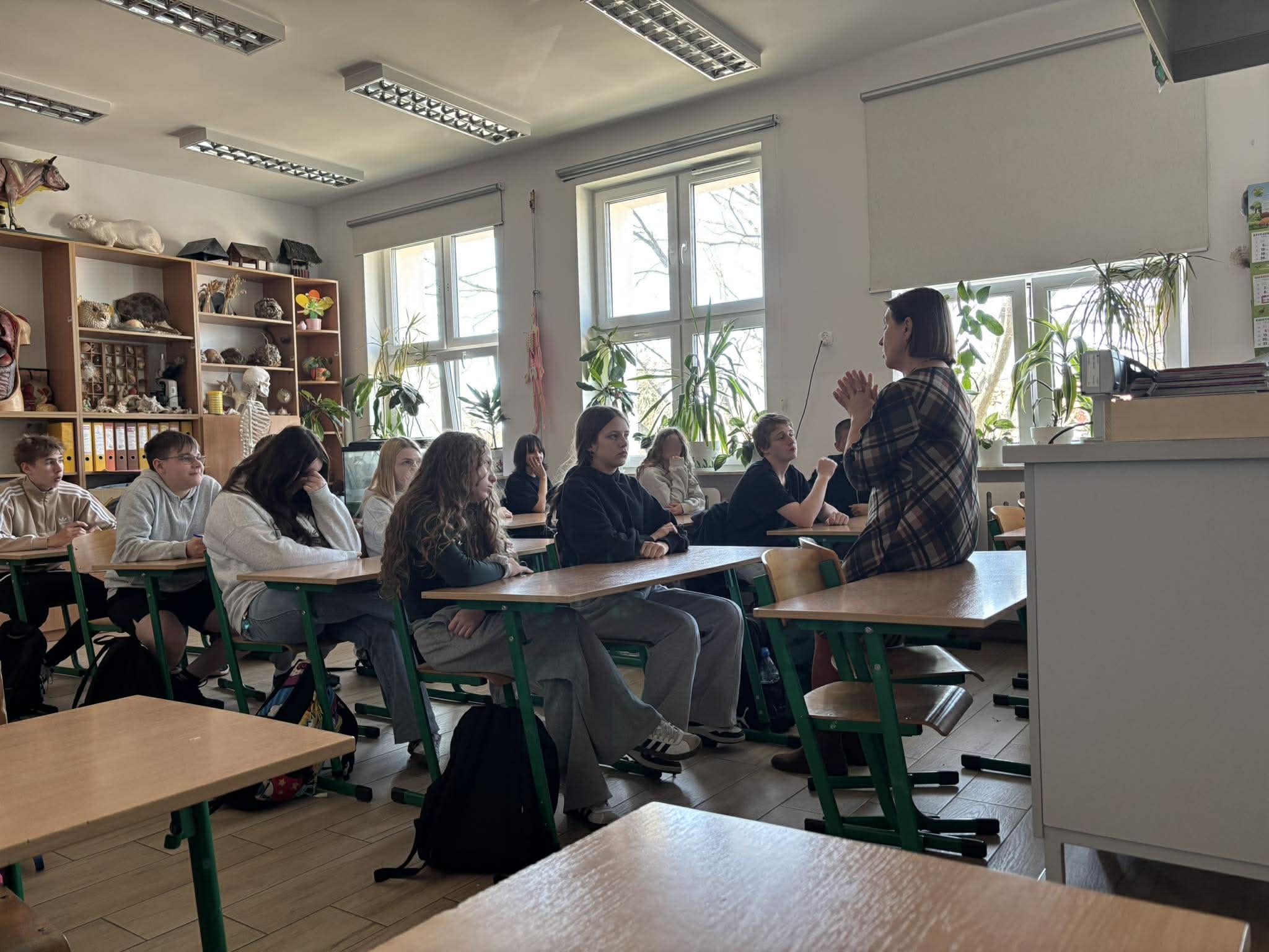 Grupa nastolatków siedzi w ławkach i uważnie słucha kobiety prowadzącej warsztaty. Prelegentka siedzi na krawędzi stołu przodem do uczniów, gestykulując dłońmi. W tle widać okna z roślinami doniczkowymi i półki z pomocami dydaktycznymi.
