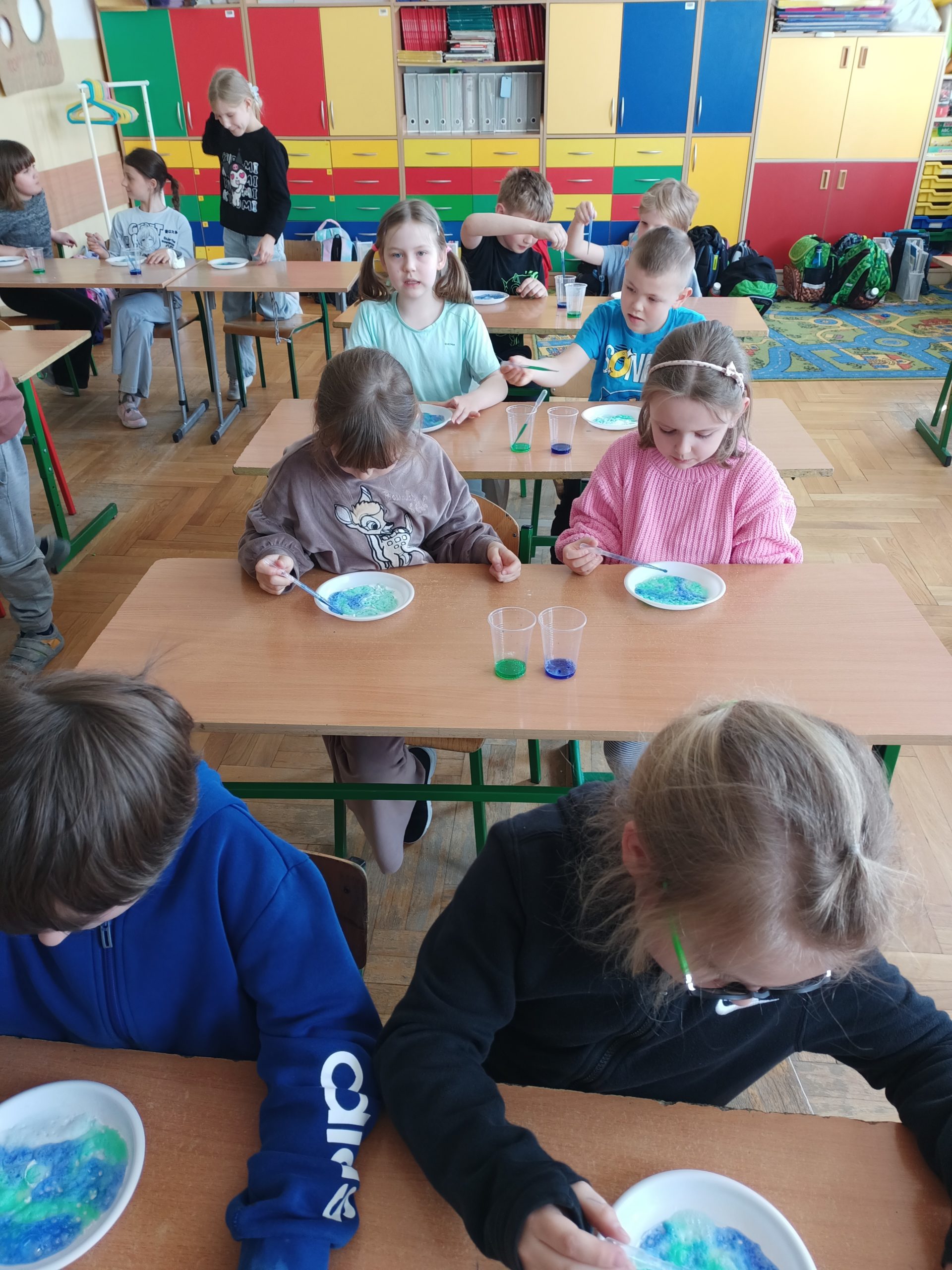 Grupa dzieci siedzących w ławkach tworzy prace plastyczne z wykorzystaniem pipet, zabarwionego octu i sody. Na każdej ławce przed dzieckiem leży talerzyk z zabarwioną sodą i stoją dwa kubeczki z niebieskim i zielonym płynem. W tle kolorowe szafki.
