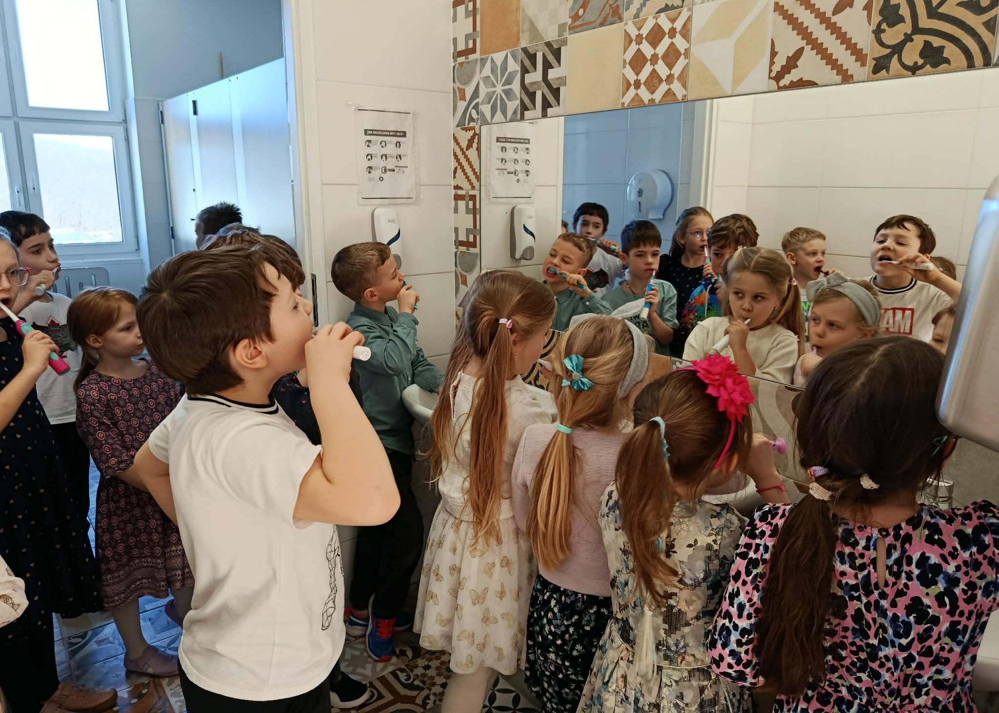 W szkolnej toalecie na tle lustra grupa dzieci szczotkuje zęby.
