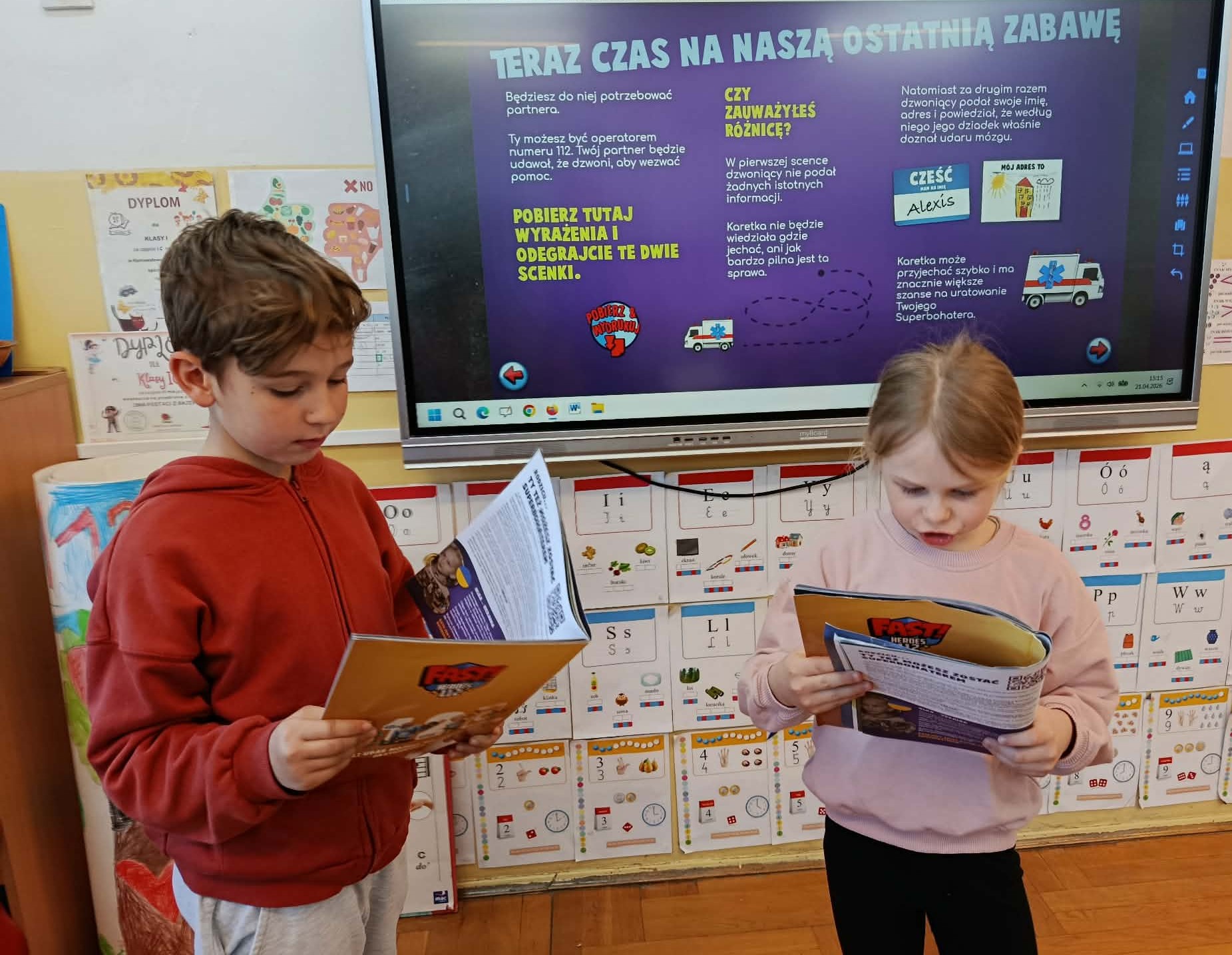 Na tle tablicy multimedialnej wyświetlającej slajd z projektu FAST Heroes stoi para uczniów z zeszytami ćwiczeń projektu FAST Heroes w rękach.