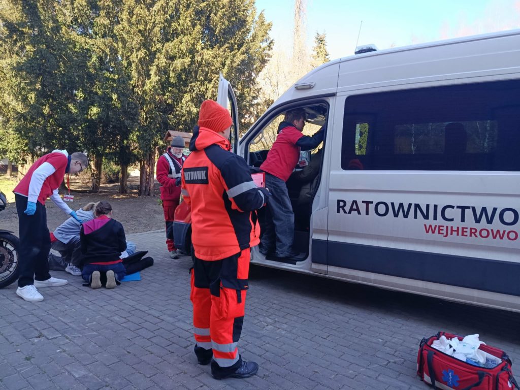 Na chodniku bus, obok motocykl i leżący motocyklista. W busie uczeń udzielający pomocy kierowcy. Obok busa trzech uczniów udzielających pomocy motocykliście. Przed nimi ratownik oceniający ich działania i apteczka.