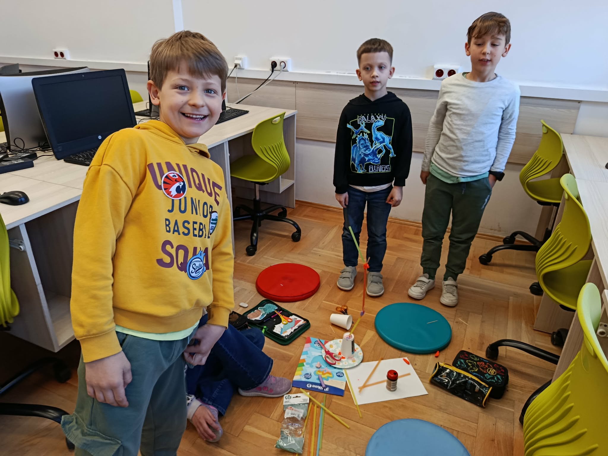 Na podłodze stoi konstrukcja ze słomek, kubeczka i plasteliny. W koło stoją trzej chłopcy, obok nich leżą materiały.