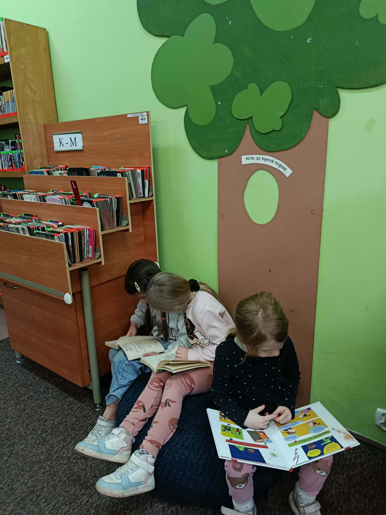 W wypożyczalni koło regału z audiobookami na pufie siedzą trzy dziewczynki z klasy 1c z książkami w dłoniach.