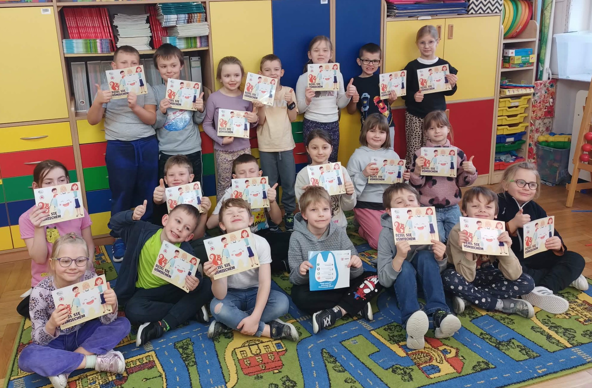 Na tle szkolnych szafek grupa dzieci trzymająca w rękach książeczki z programu „Dziel się uśmiechem”.
