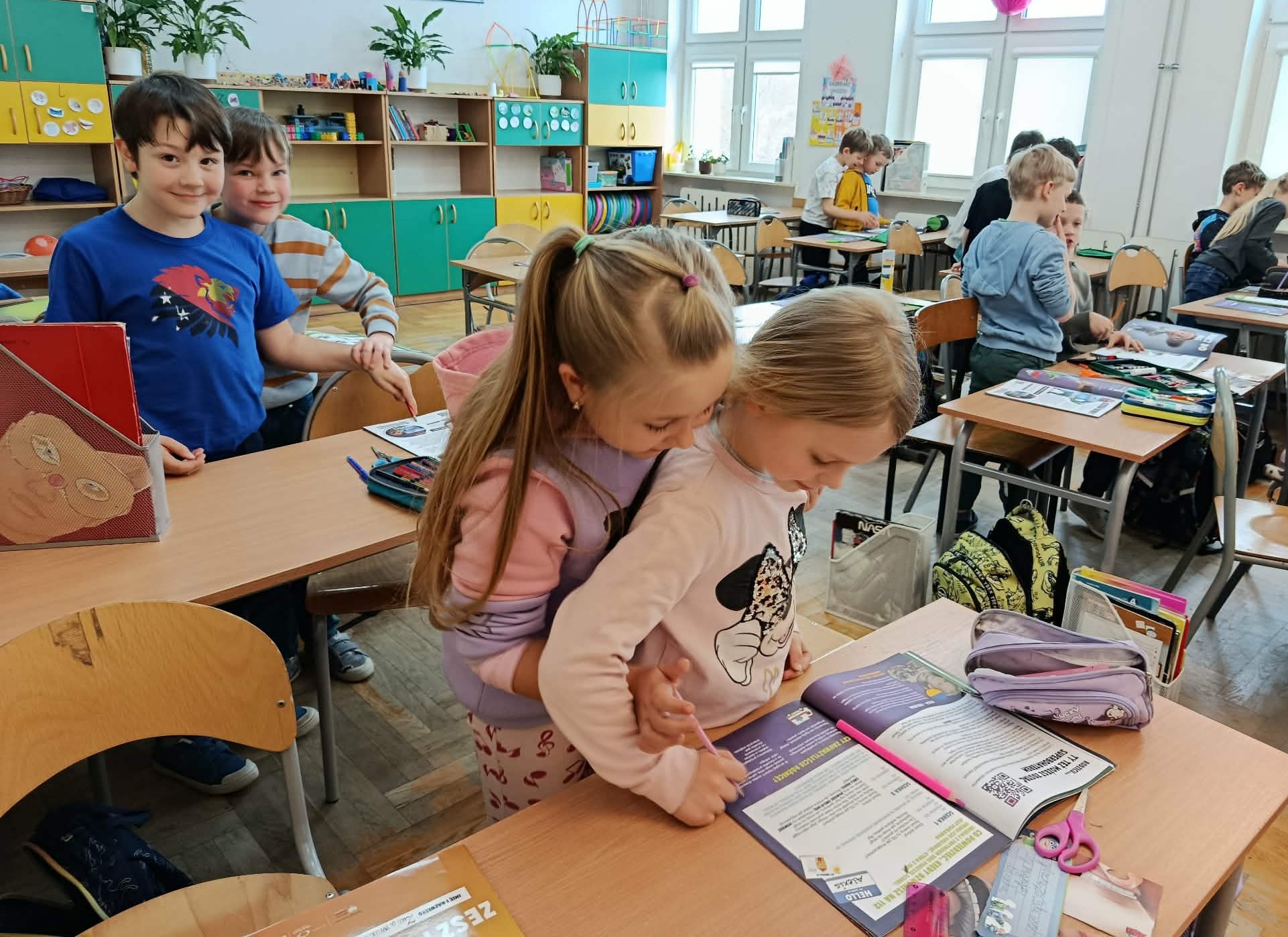 Na pierwszym planie uczennica stojąca za koleżanką przy ławce i próbująca pisać w ćwiczeniach ręką koleżanki. Za nimi para uczniów i reszta klasy.