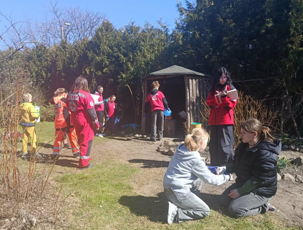 W ogrodzie wiata. Przed nią drużyna udzielająca pomocy poszkodowanym. Obok ratownicy oceniających ich działania.