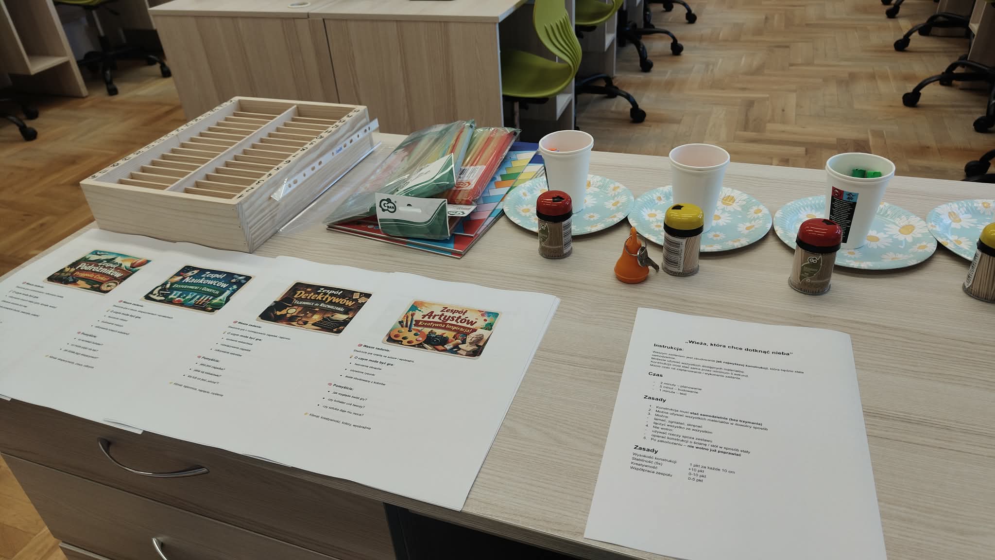 Na biurku przygotowane kartki z zadaniami dla grup, obok materiały: słomki, kubeczki, ta-lerzyki, wykałaczki.