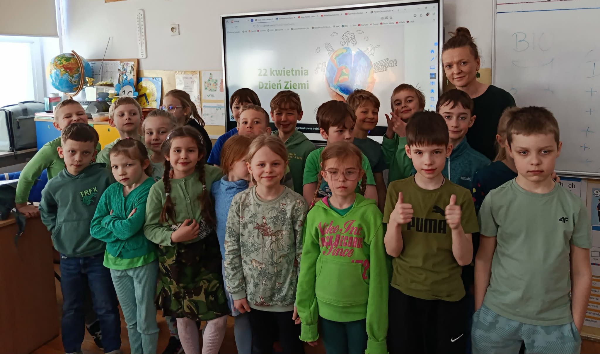 Grupa uśmiechniętych dzieci w zielonych ubraniach pozuje razem z nauczycielką na tle białej tablicy w sali lekcyjnej.