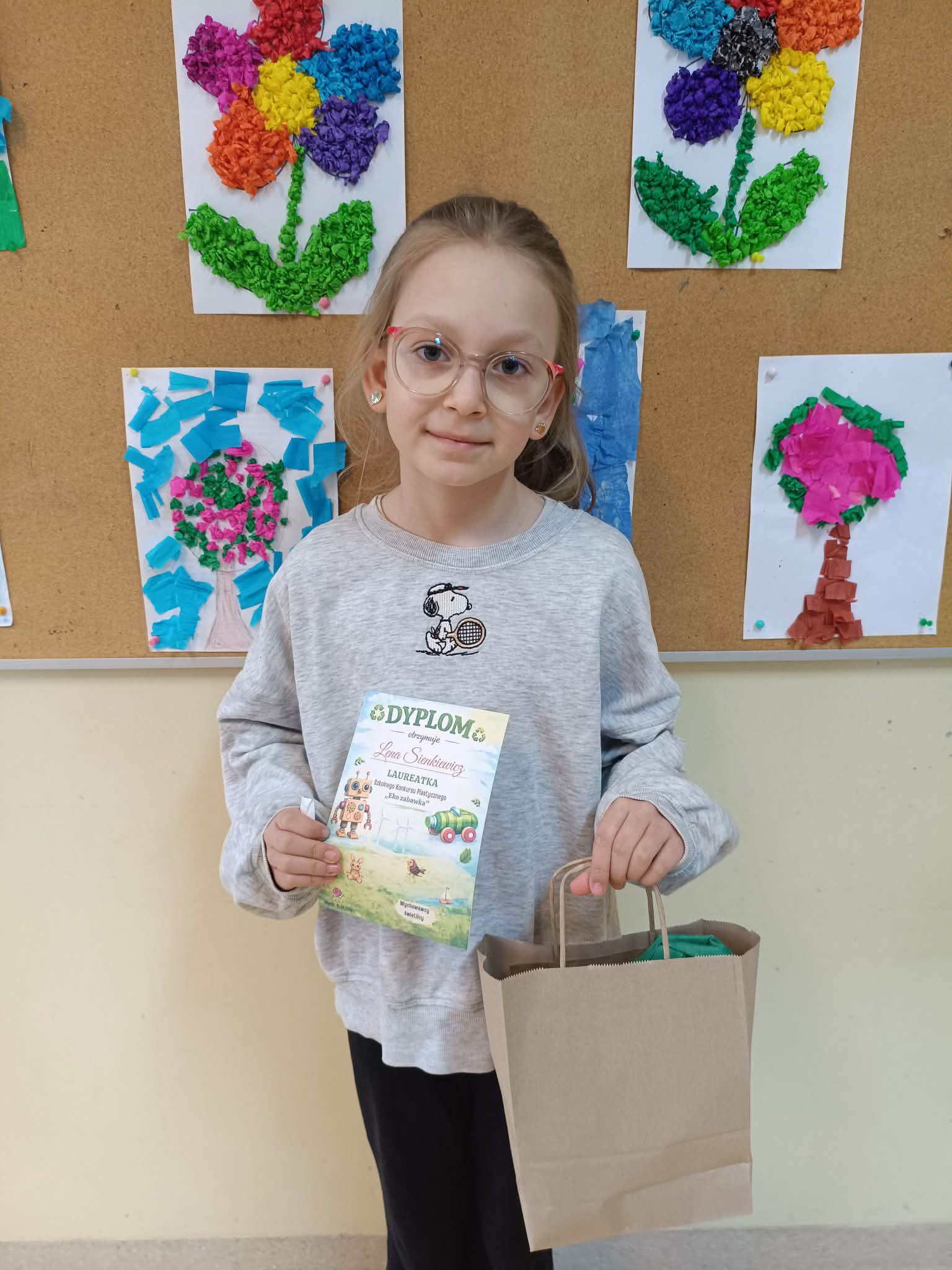 Dziewczynka stoi w sali, trzyma dyplom i papierowa torbe, na tle tablicy z pracami plastycznymi przedstawiającymi kwiaty.