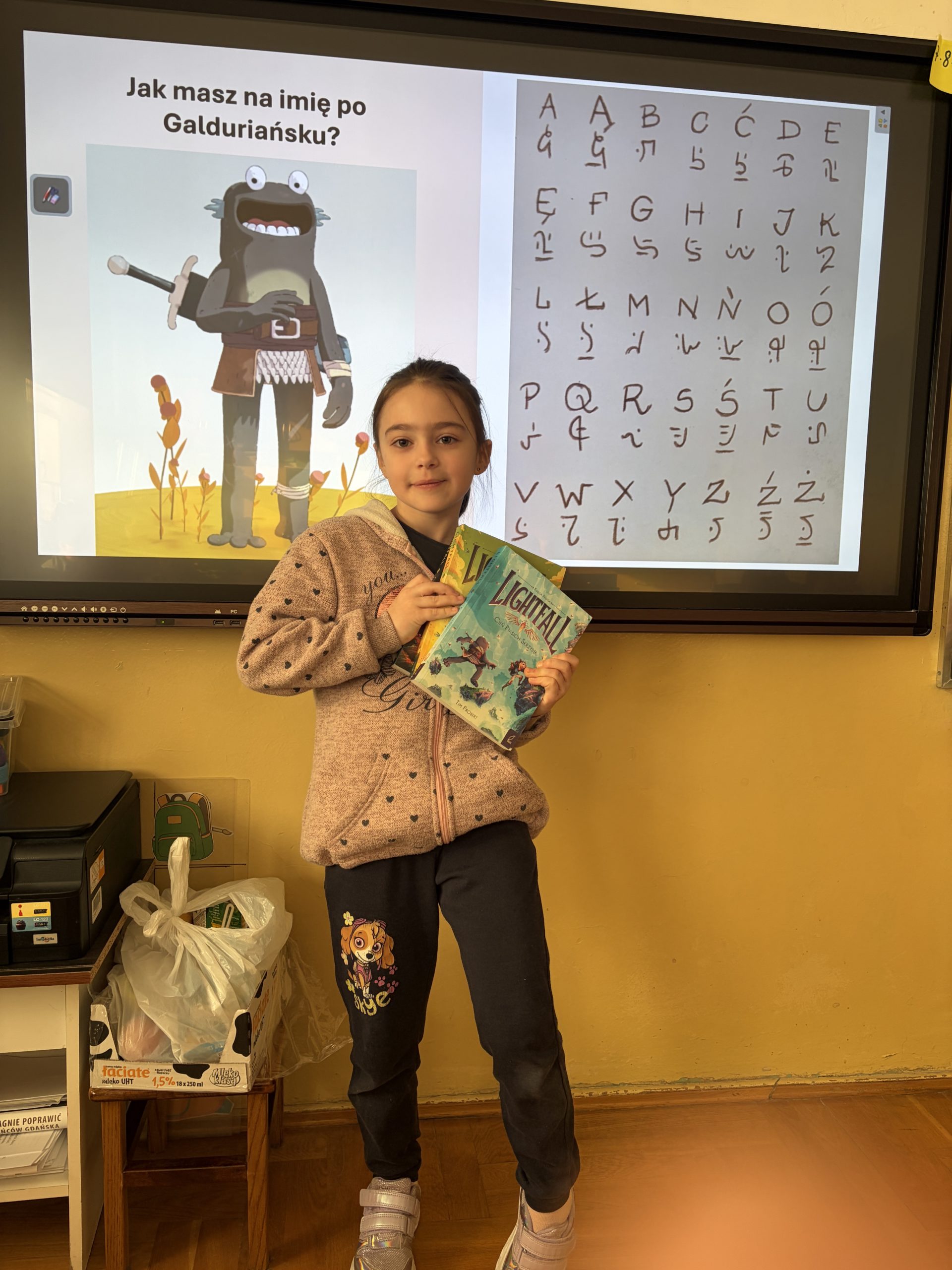 Dziewczynka w beżowej bluzie z kapturem stoi przed interaktywnym ekranem, trzymając w rękach dwie książki z serii „Lightfall”, podczas gdy na ekranie za nią widnieje prezentacja z postacią z bajki i alfabetem.