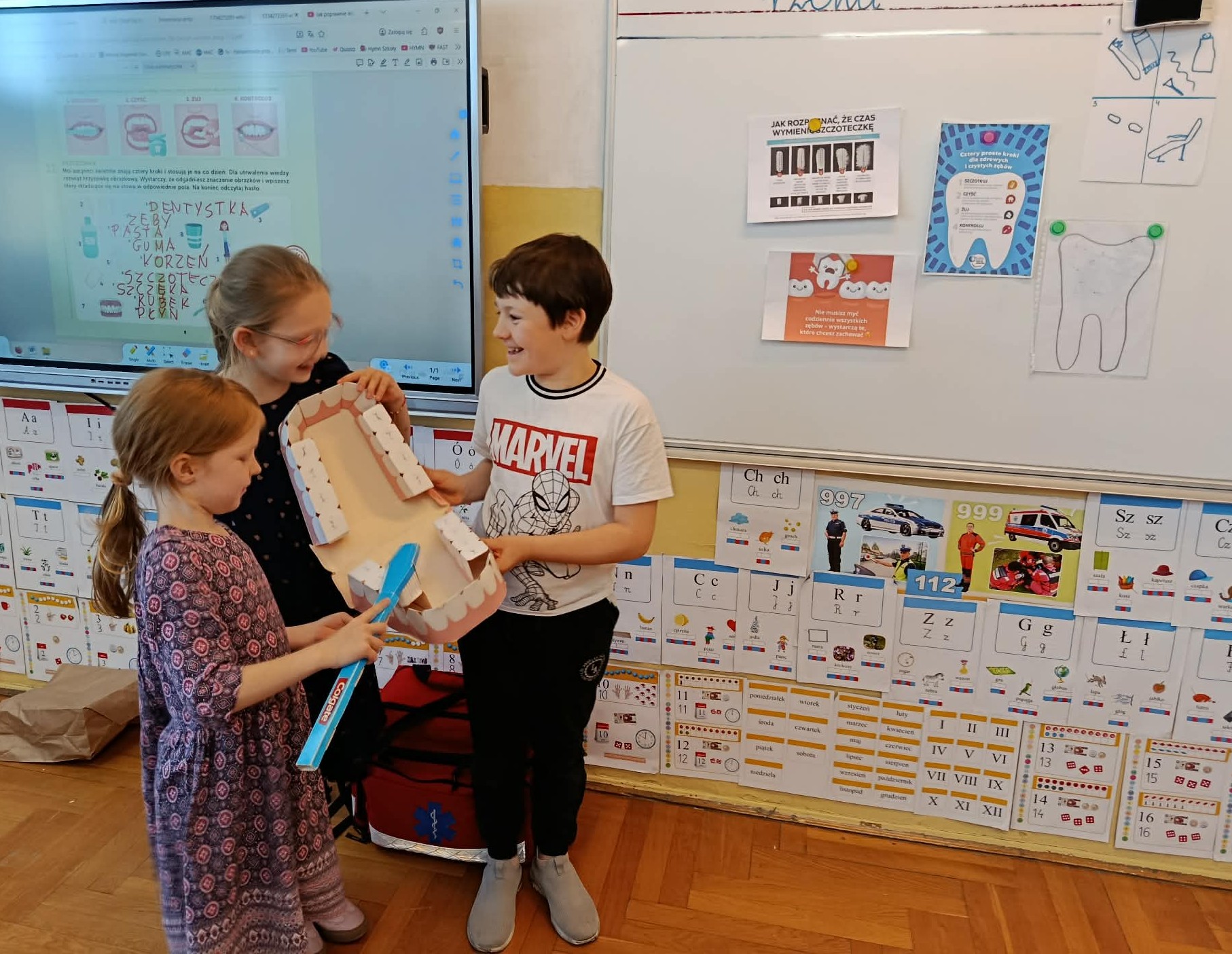 Na tle tablicy i ekranu multimedialnego z wyświetlaną krzyżówką troje dzieci trzymający model szczęki i szczotkujący zęby.